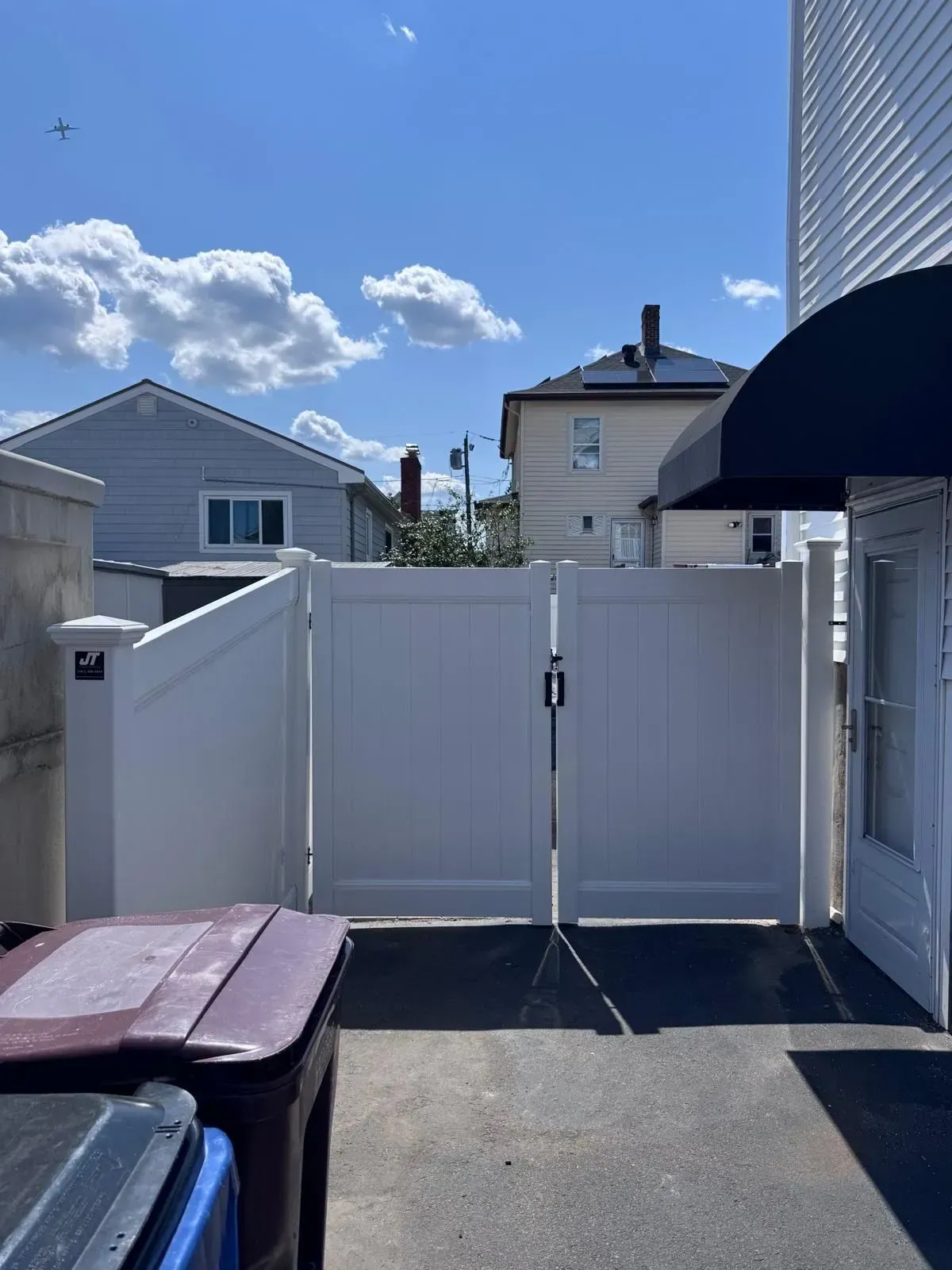 Backyard with white fence gate, blue sky, neighboring houses, and trash bins in the foreground.