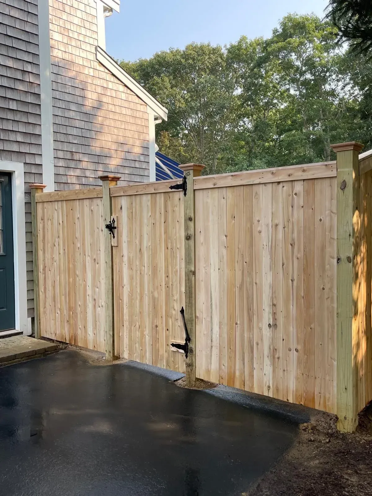 Wooden driveway gate beside a brick house, with wet pavement and trees in the background.