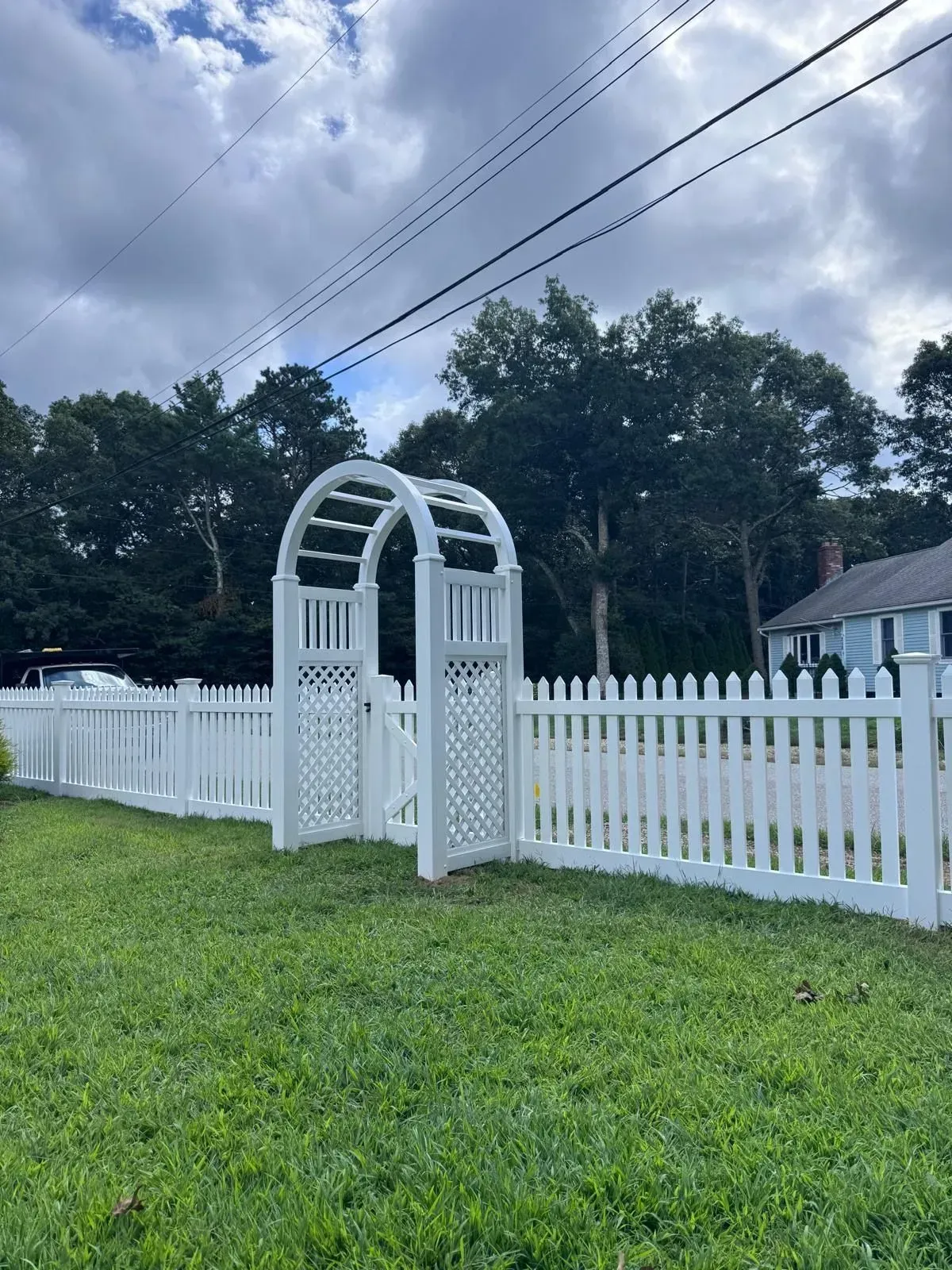 White picket fence with arched gate in a grassy yard under a cloudy sky.