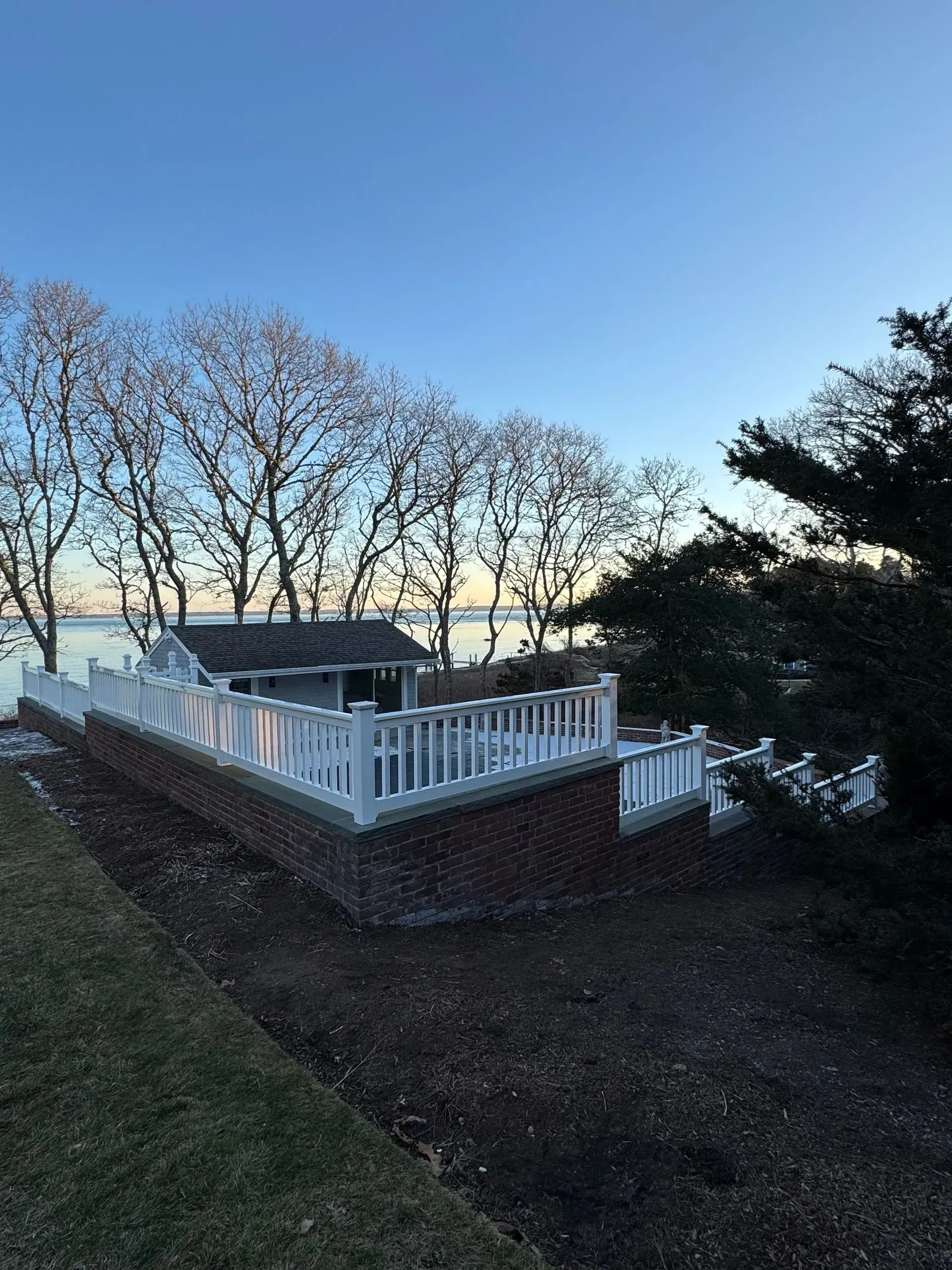 White fence and brick patio overlook a wooded landscape at dusk.