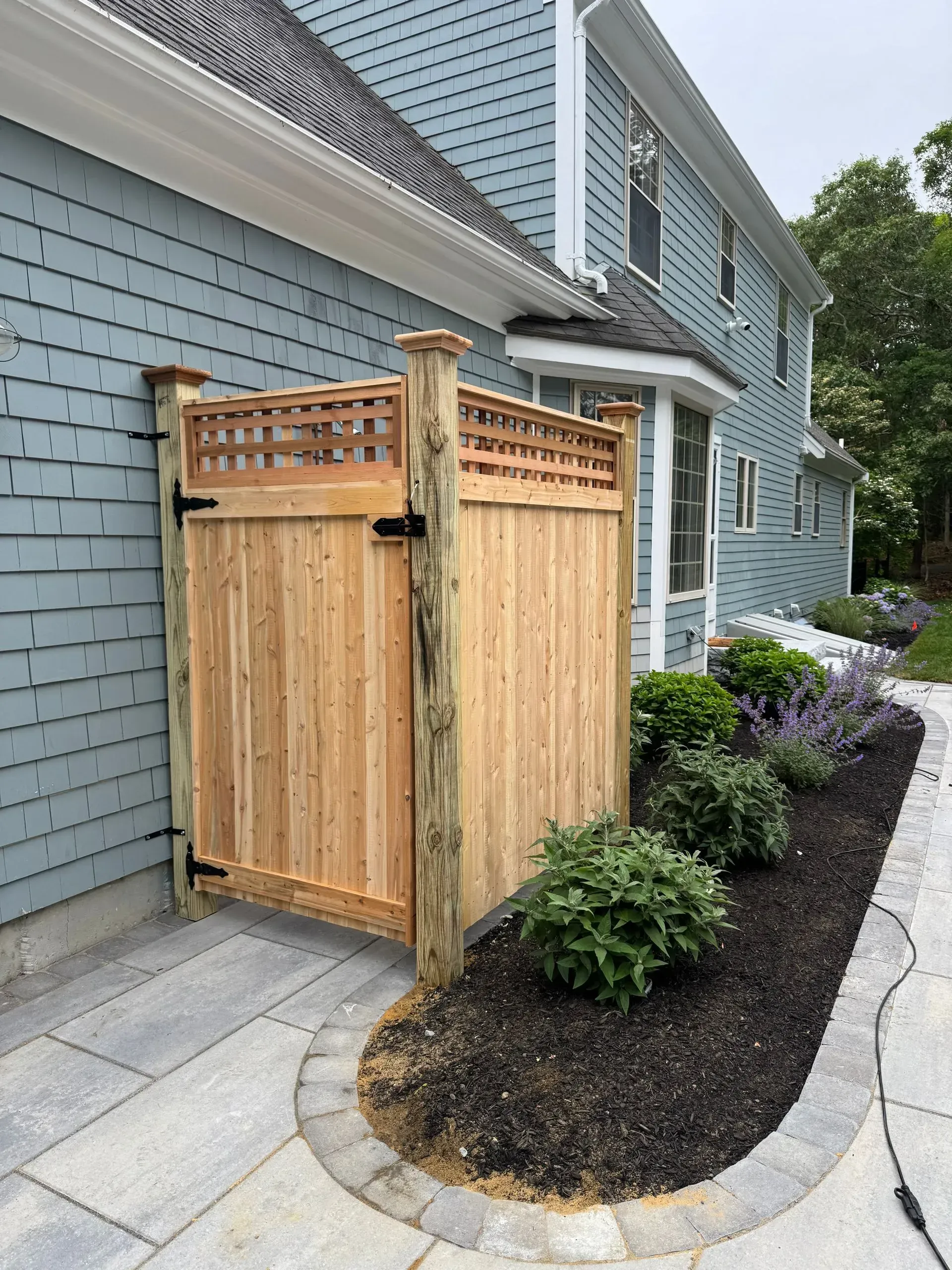 Wooden outdoor shower enclosure beside a blue house and landscaped garden path.