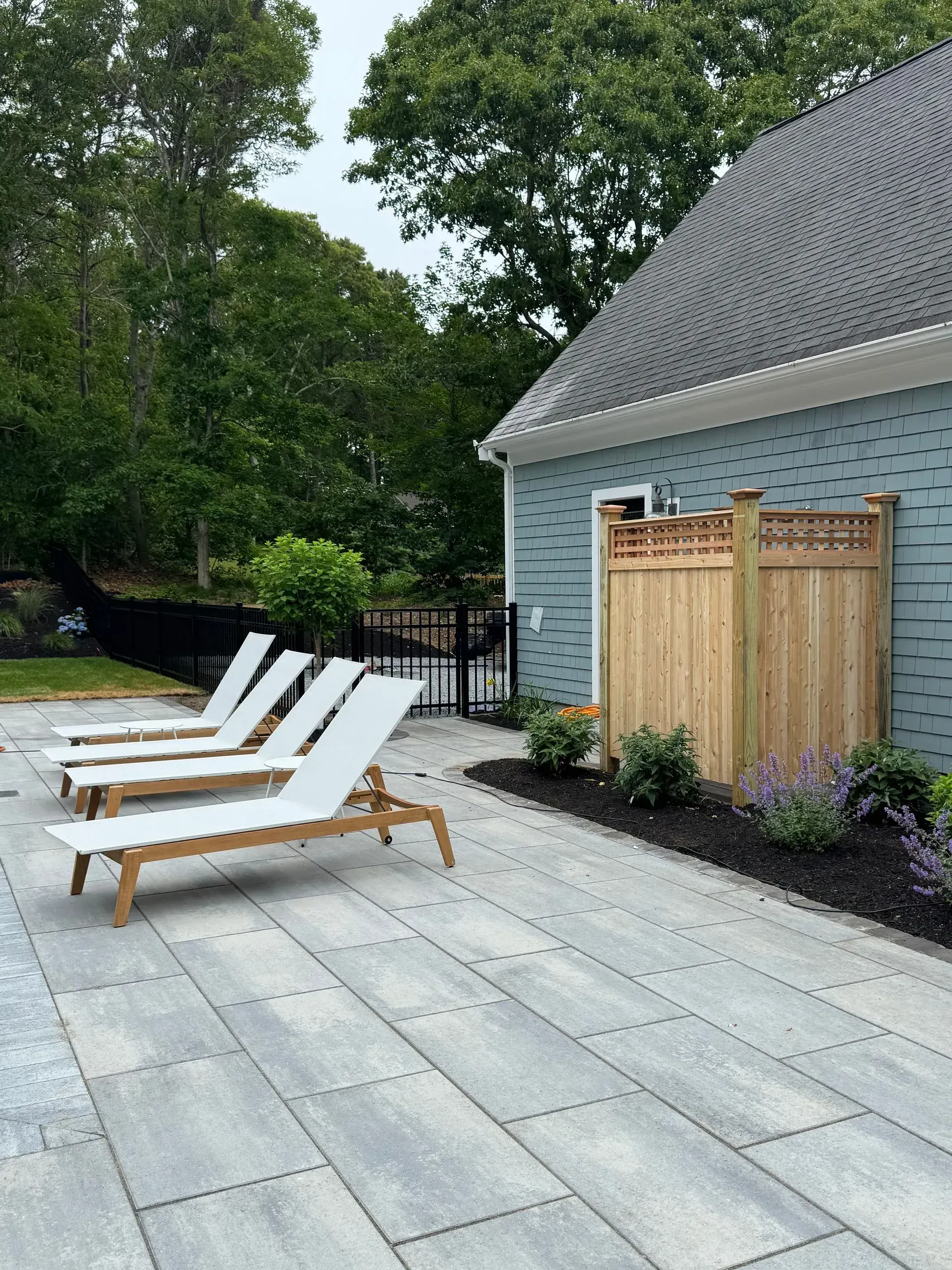 Backyard patio with three white lounge chairs, a wooden fence, and a gray-roofed house amid trees.