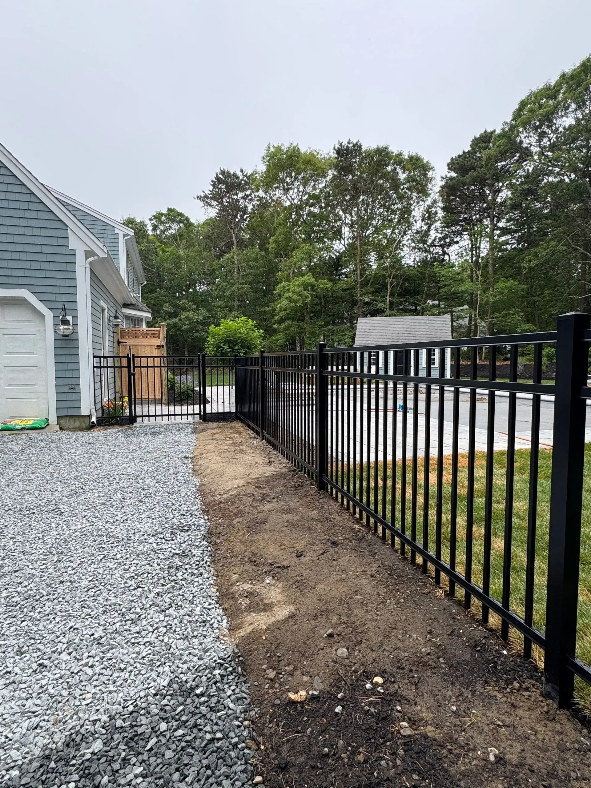 Gravel side yard beside a gray house with black metal fence and trees beyond.