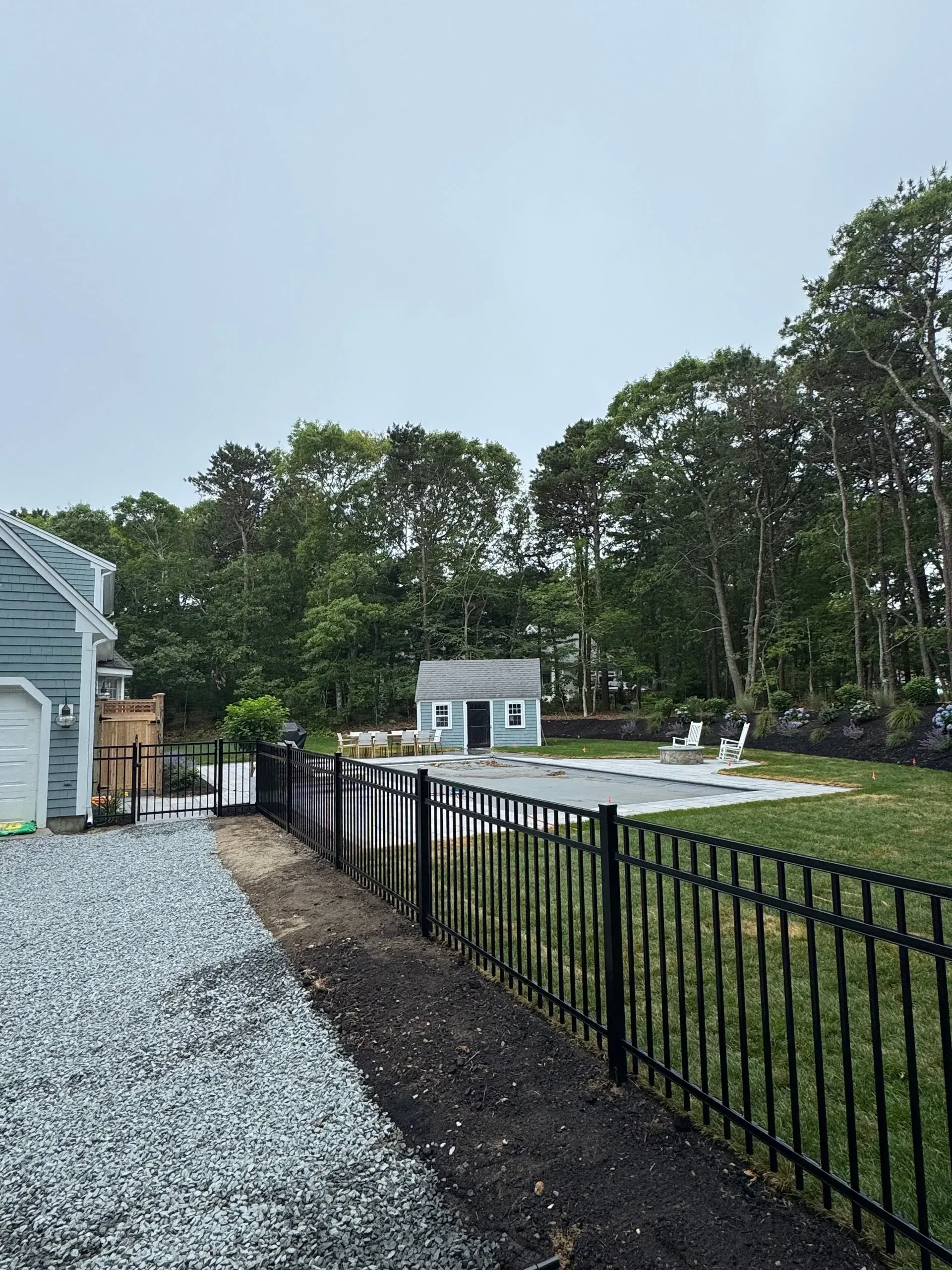 Backyard with black metal fence, gravel path, small shed, and trees under a cloudy sky.