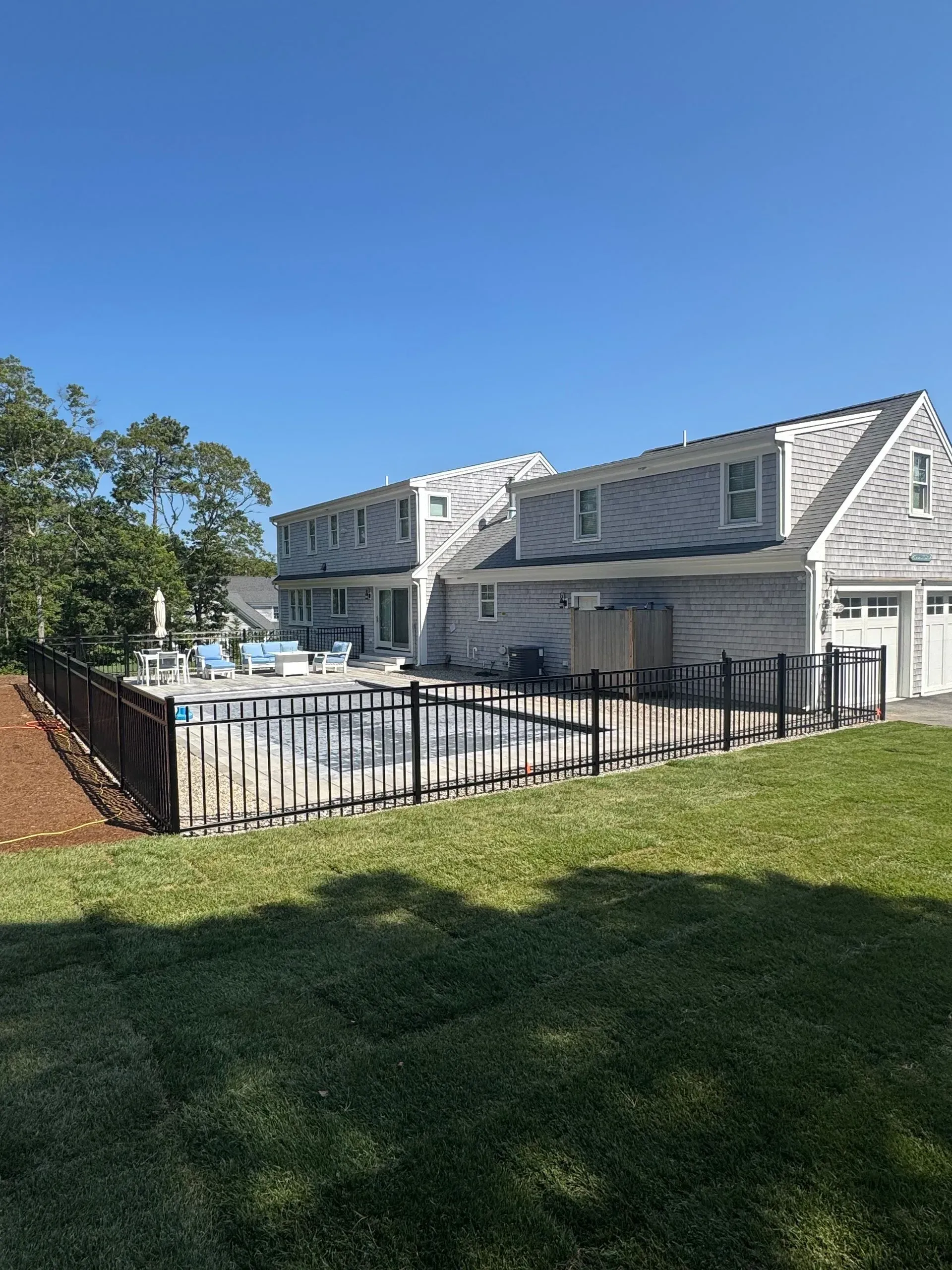 Gray building with a fenced outdoor pool deck beside a grassy lawn under a blue sky.