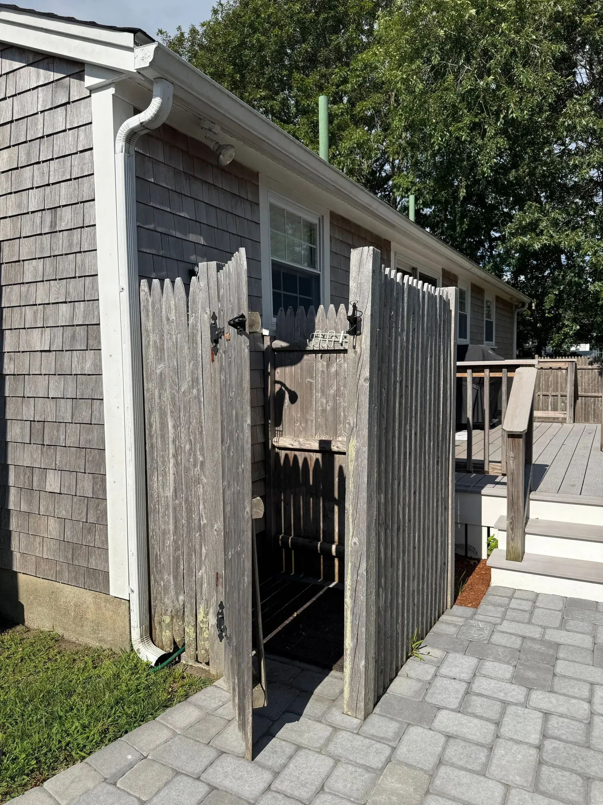 Open wooden gate beside a gray shingled house, leading to a paved walkway and patio.