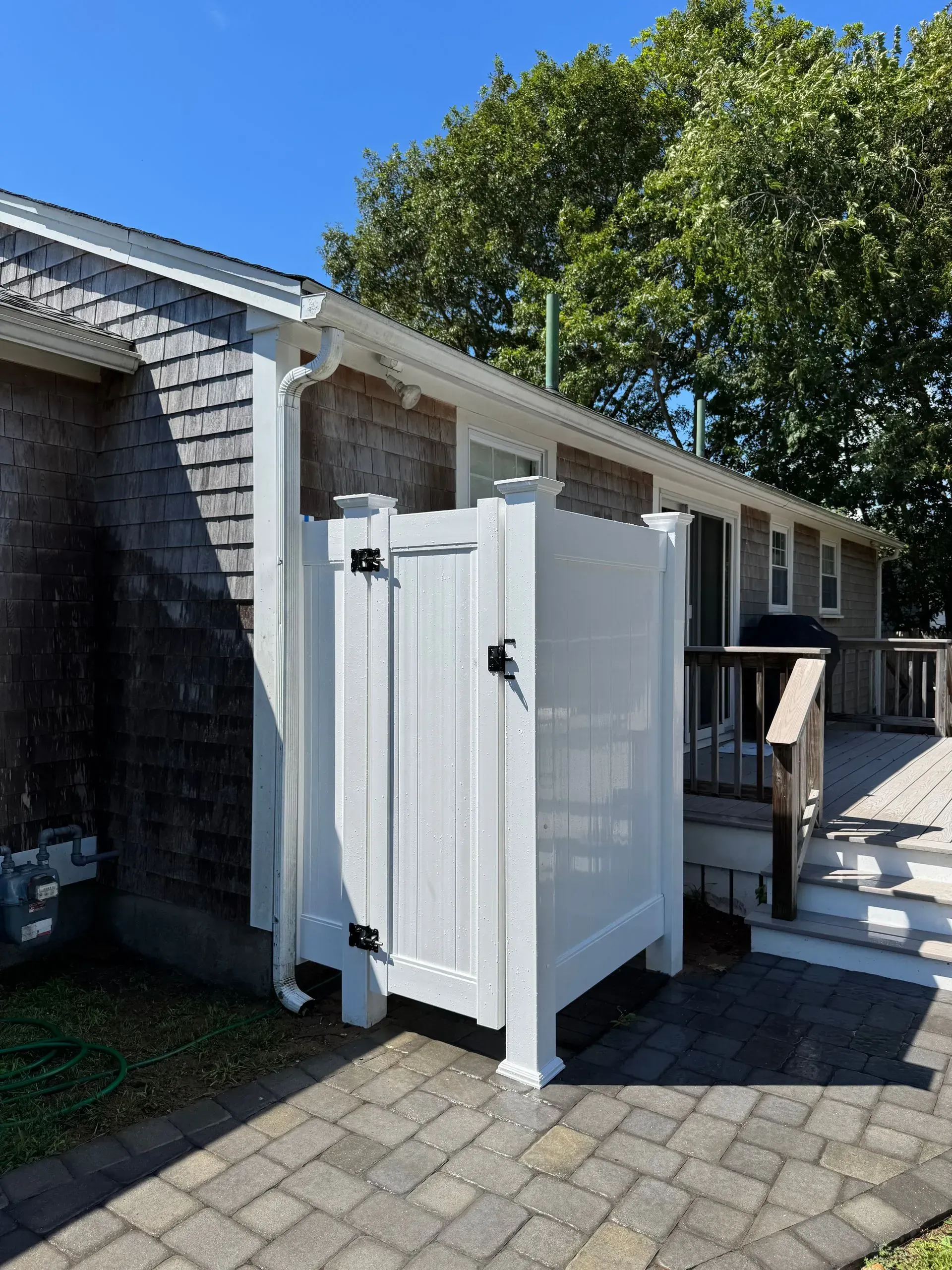 White outdoor shower stall beside a house with white trim and paving stones under a tree.