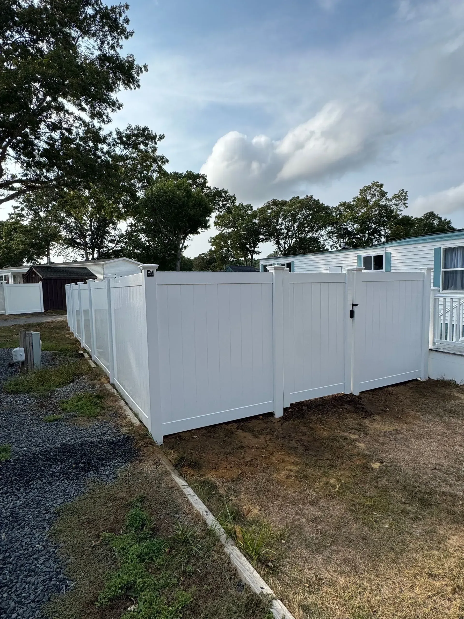 White fenced outdoor area beside a mobile home, with gravel, grass, and trees under a cloudy sky.