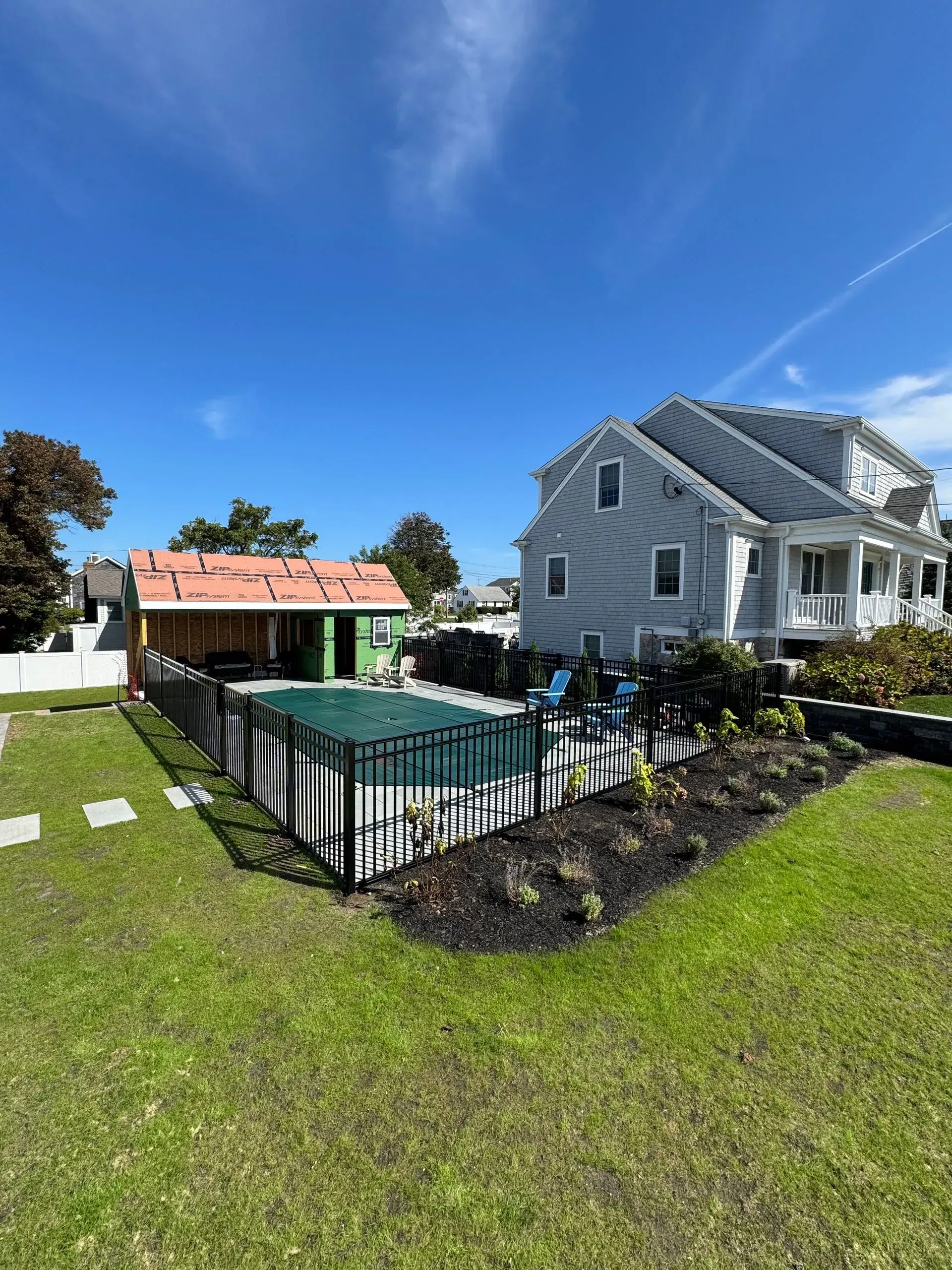 Backyard with a fenced pool, gray house, and red-roofed patio under a clear blue sky.