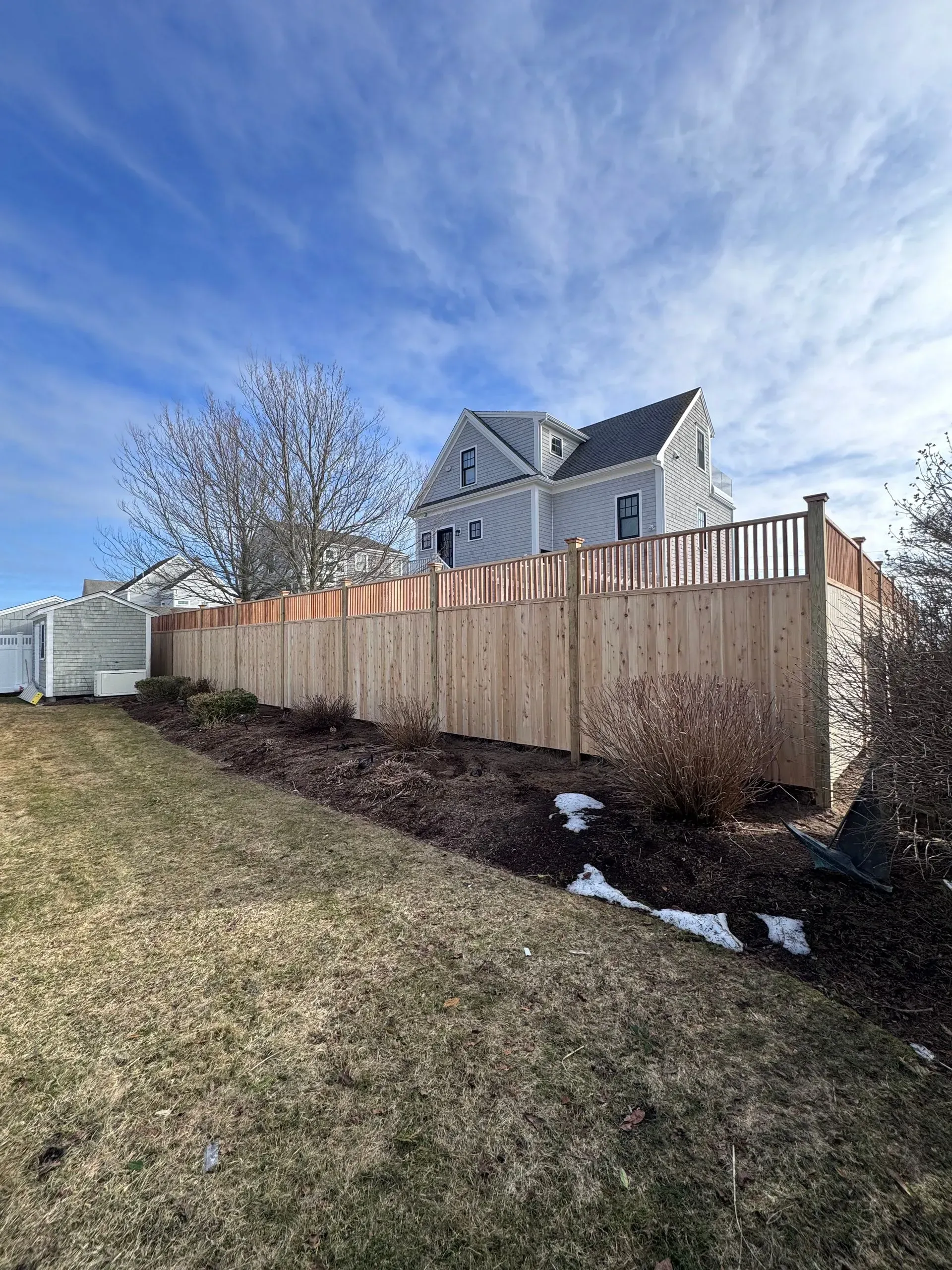 Wooden fence along a sloped yard with a gray house and blue sky in the background.