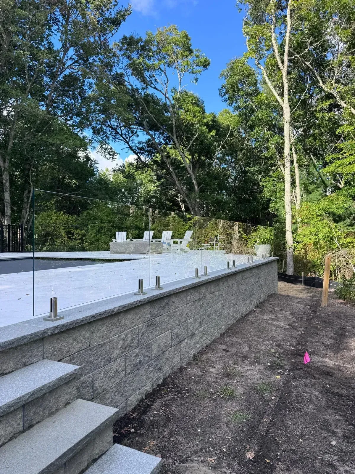 Outdoor patio with white steps, stone retaining wall, and lounge chairs beside trees and blue sky.