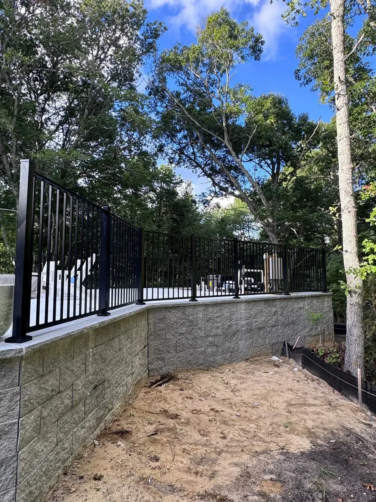 Stone retaining wall with black fence along a sandy path under trees and blue sky.