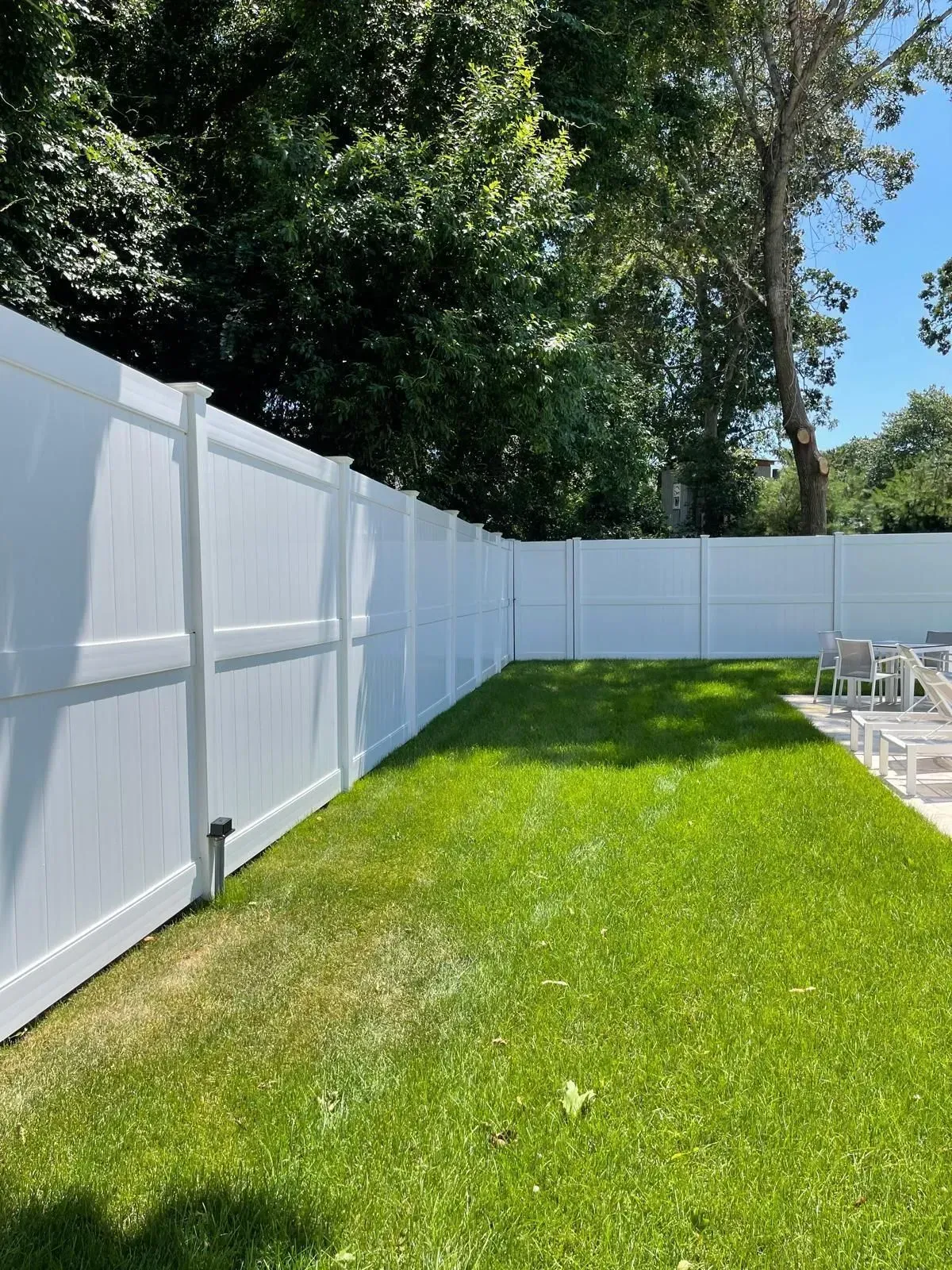 White vinyl fence bordering a sunny green backyard with trees and a patio area.