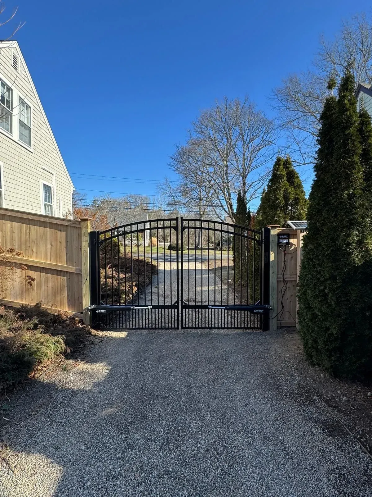 Gravel driveway leading to a black metal gate beside a house and tall hedges under a clear blue sky.
