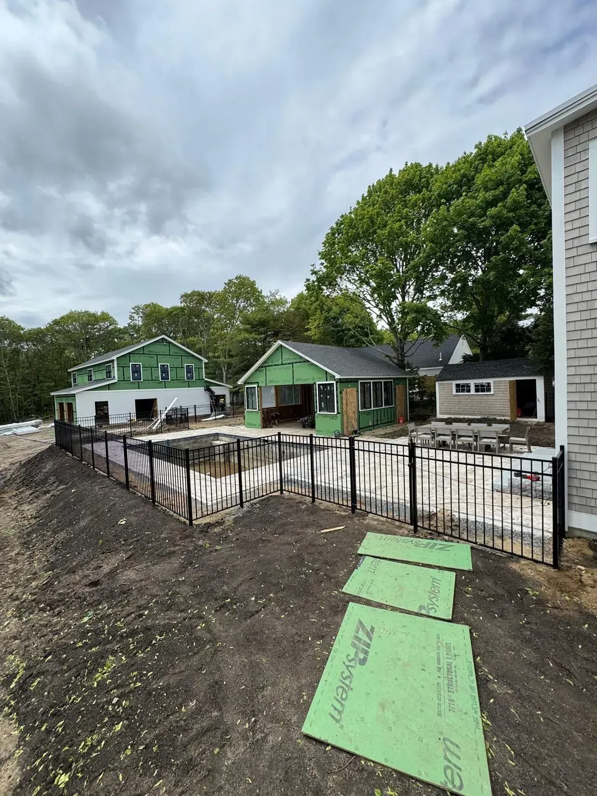Partly built houses with green siding and foundation trenches beside a fenced dirt lot under cloudy sky.