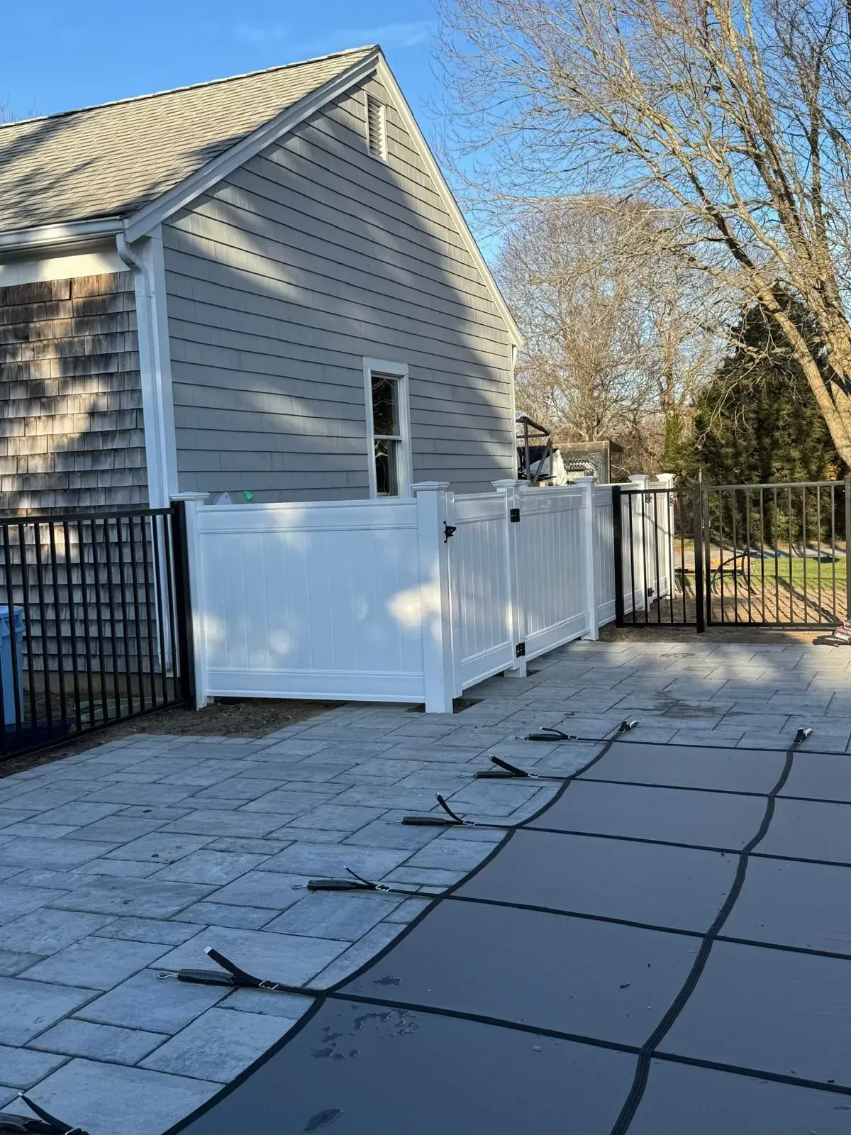 Patio beside a gray house with a white fence and black gate on a sunny day.