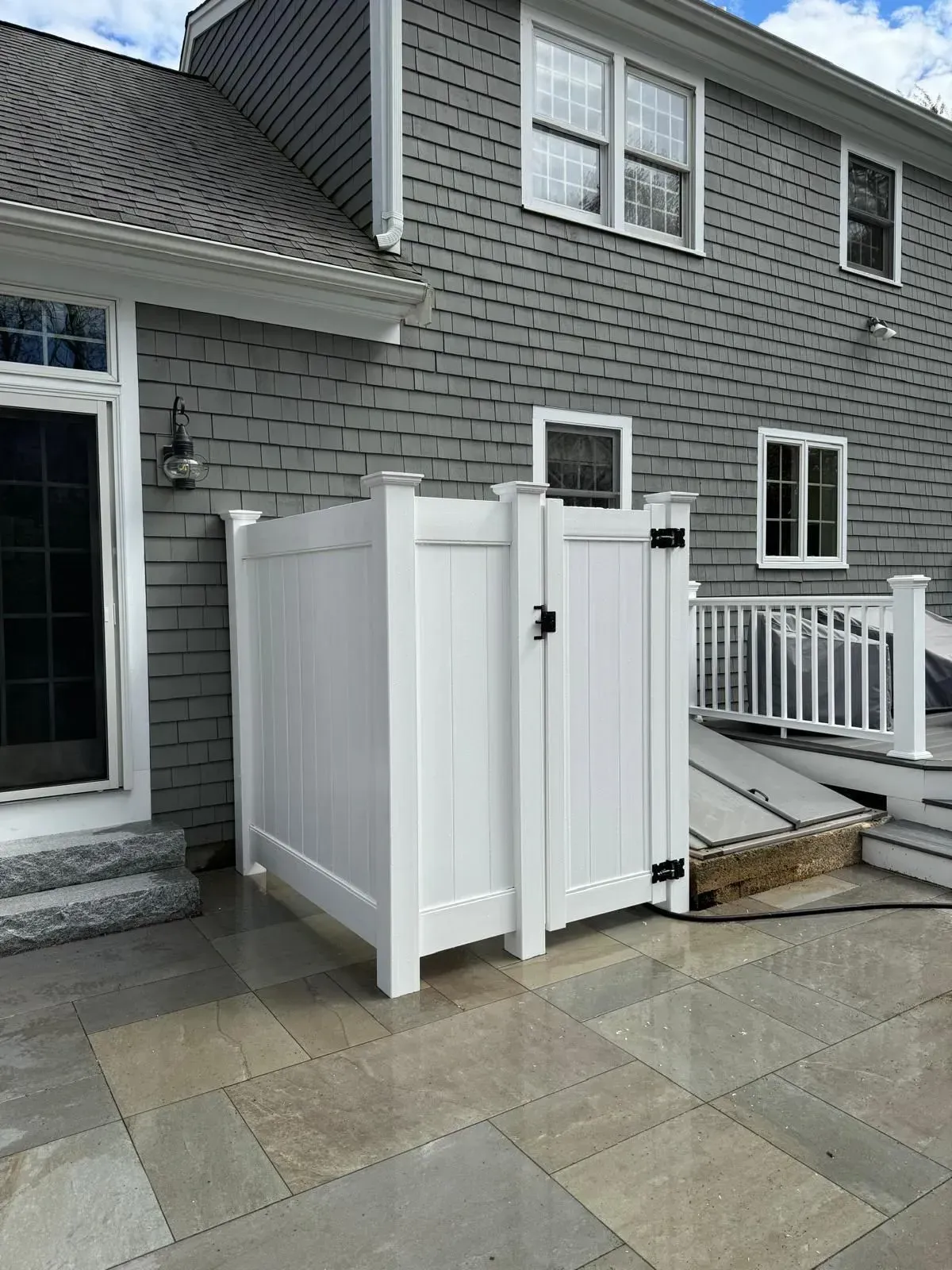 White outdoor shower enclosure on a stone patio beside a gray house with a porch and deck stairs.