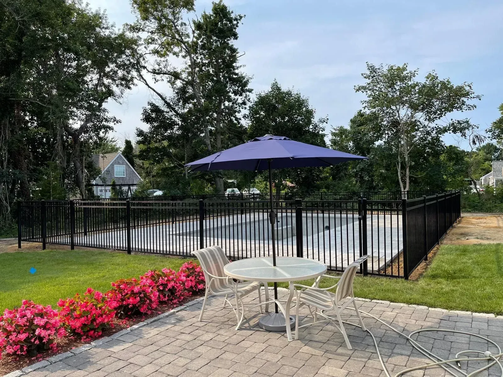 Patio table with purple umbrella by a fenced pool, bordered by pink flowers and trees.