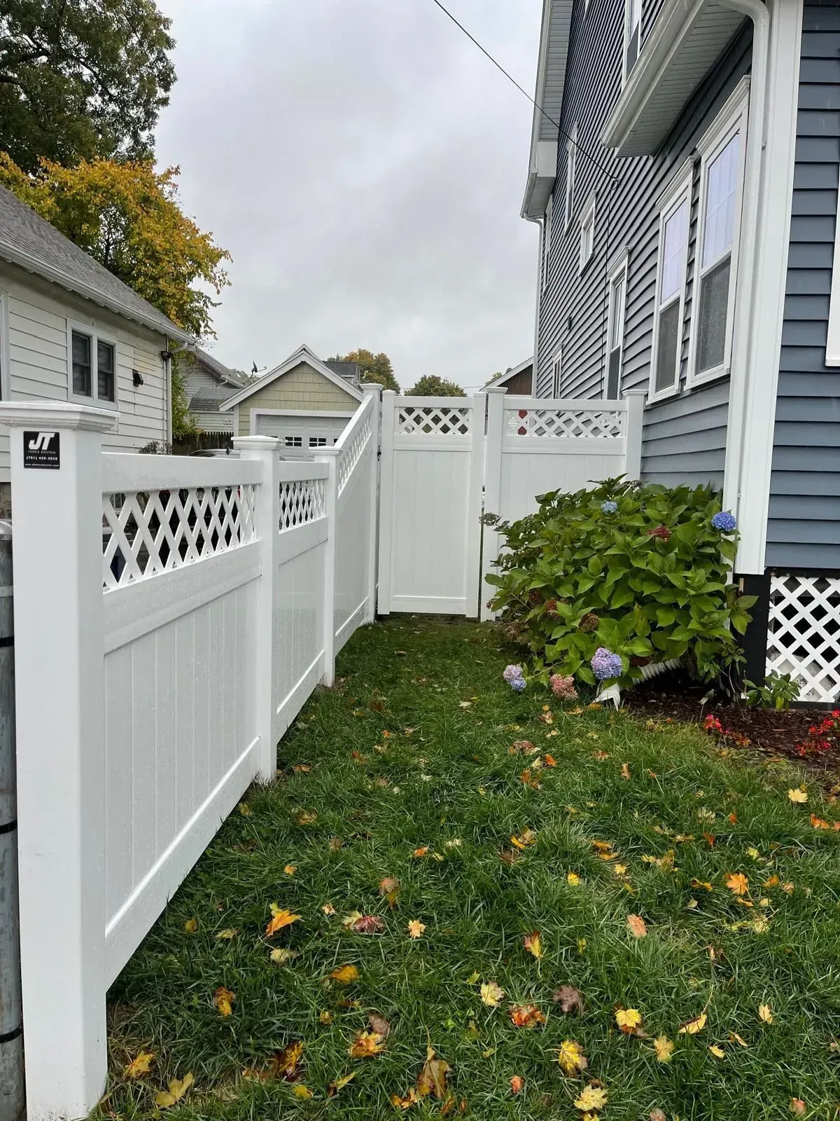 Narrow grassy side yard with white lattice fences and a blue-gray house beside a large shrub.