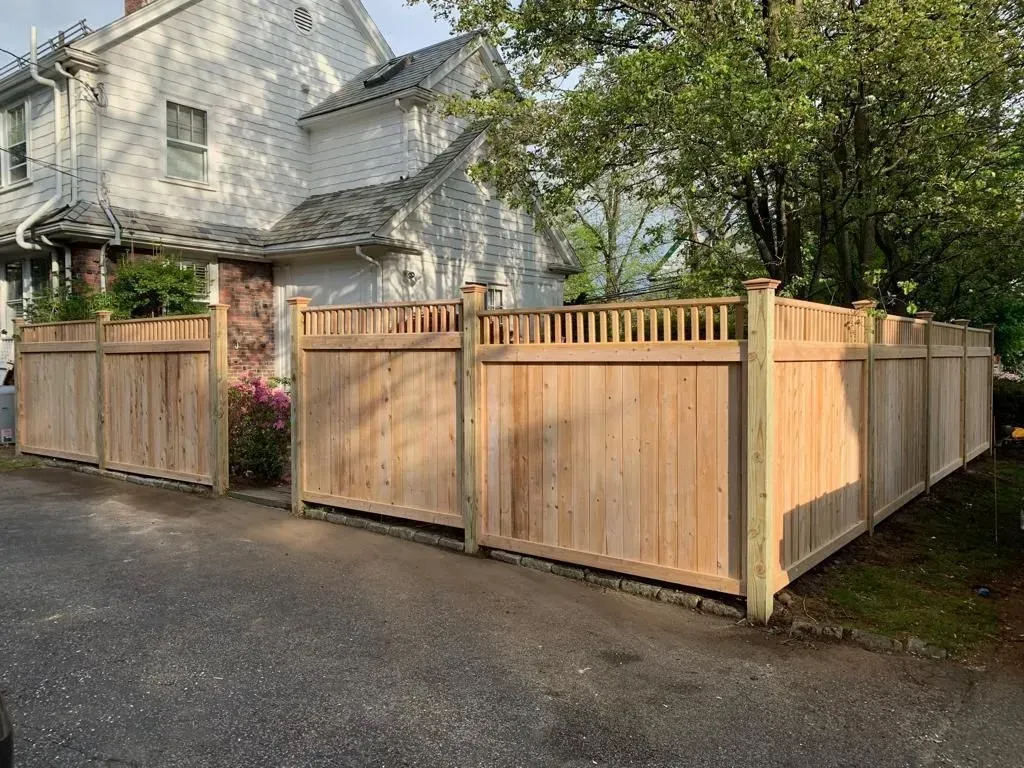 Wooden privacy fence and gates enclosing a driveway beside a white house and trees.