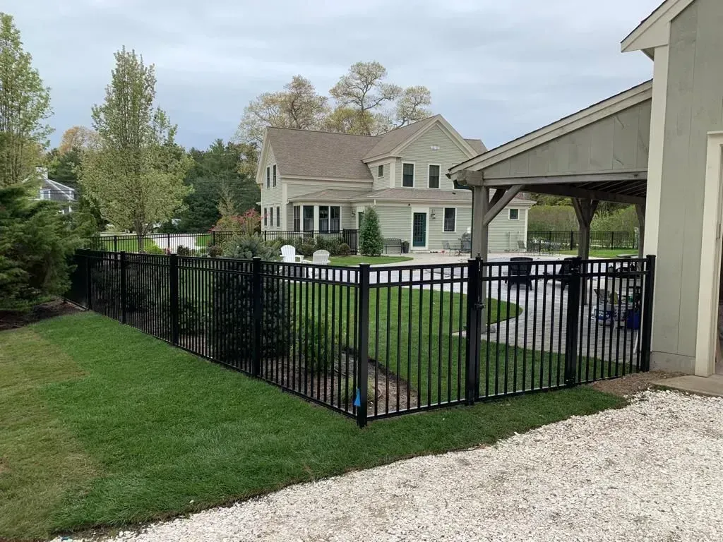 Black metal fence enclosing a green lawn beside a house and gravel driveway under an overcast sky.