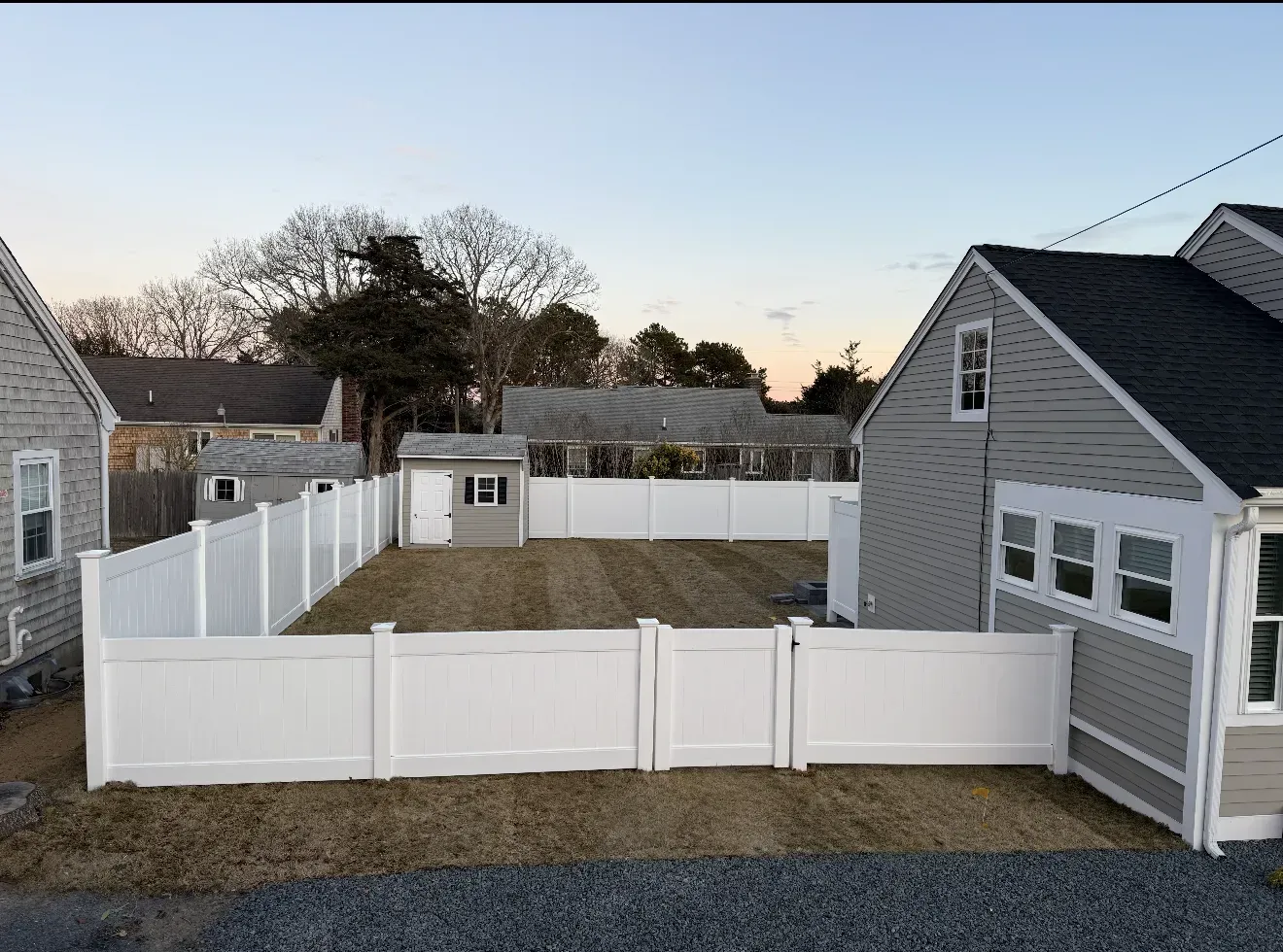White-fenced backyard with a small shed, gray house, and gravel area at dusk.
