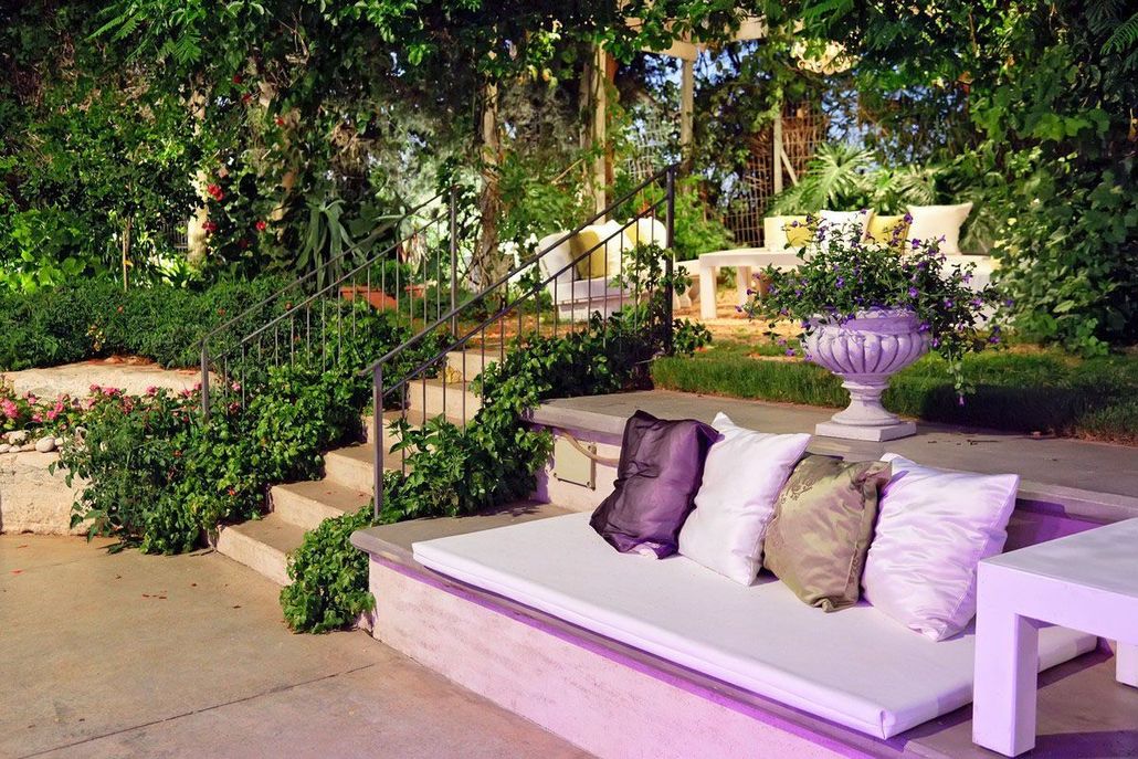 Outdoor patio with white seating, purple pillows, and lush greenery beside a staircase.