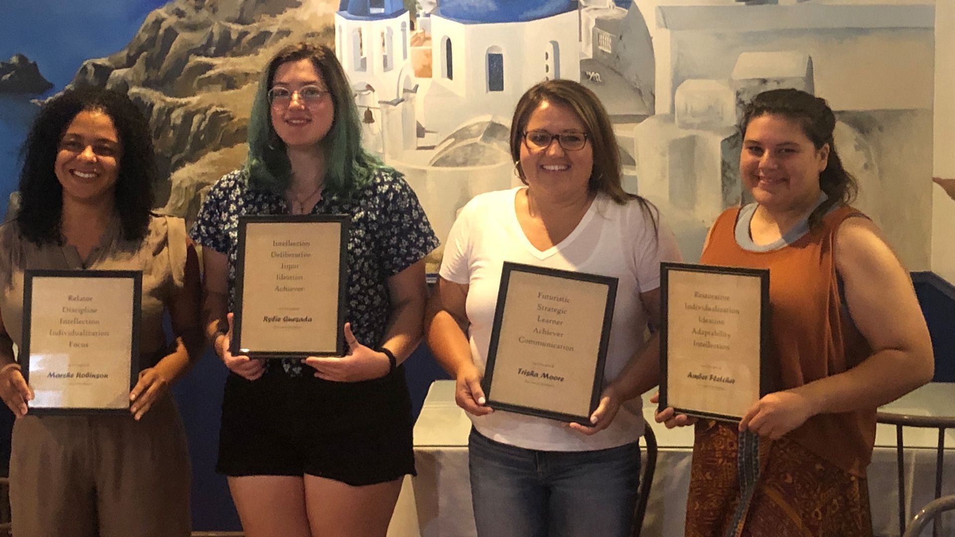 Four women holding framed awards stand together indoors, smiling in front of a painted wall.