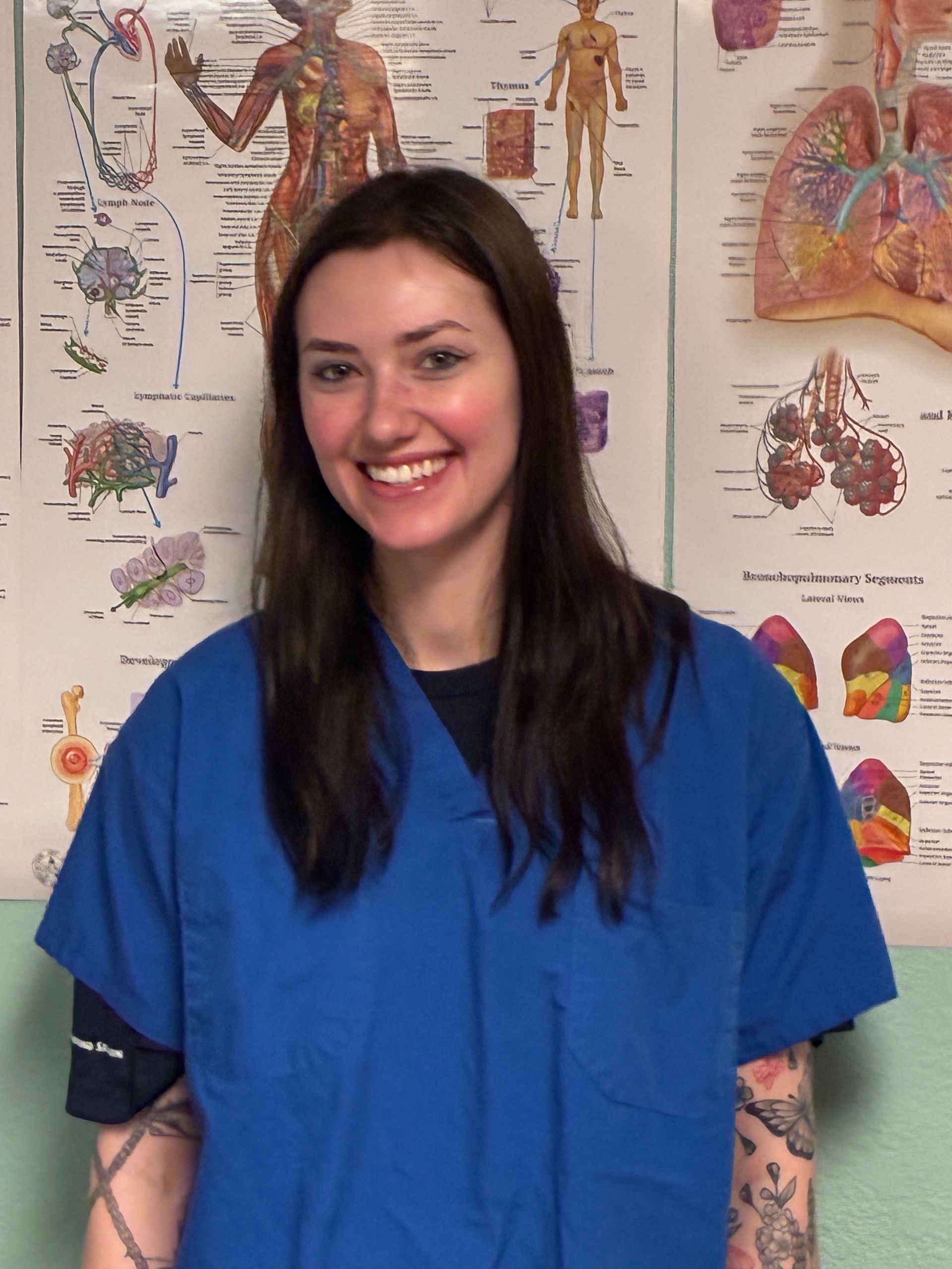 Smiling person in blue scrubs standing before medical anatomy posters