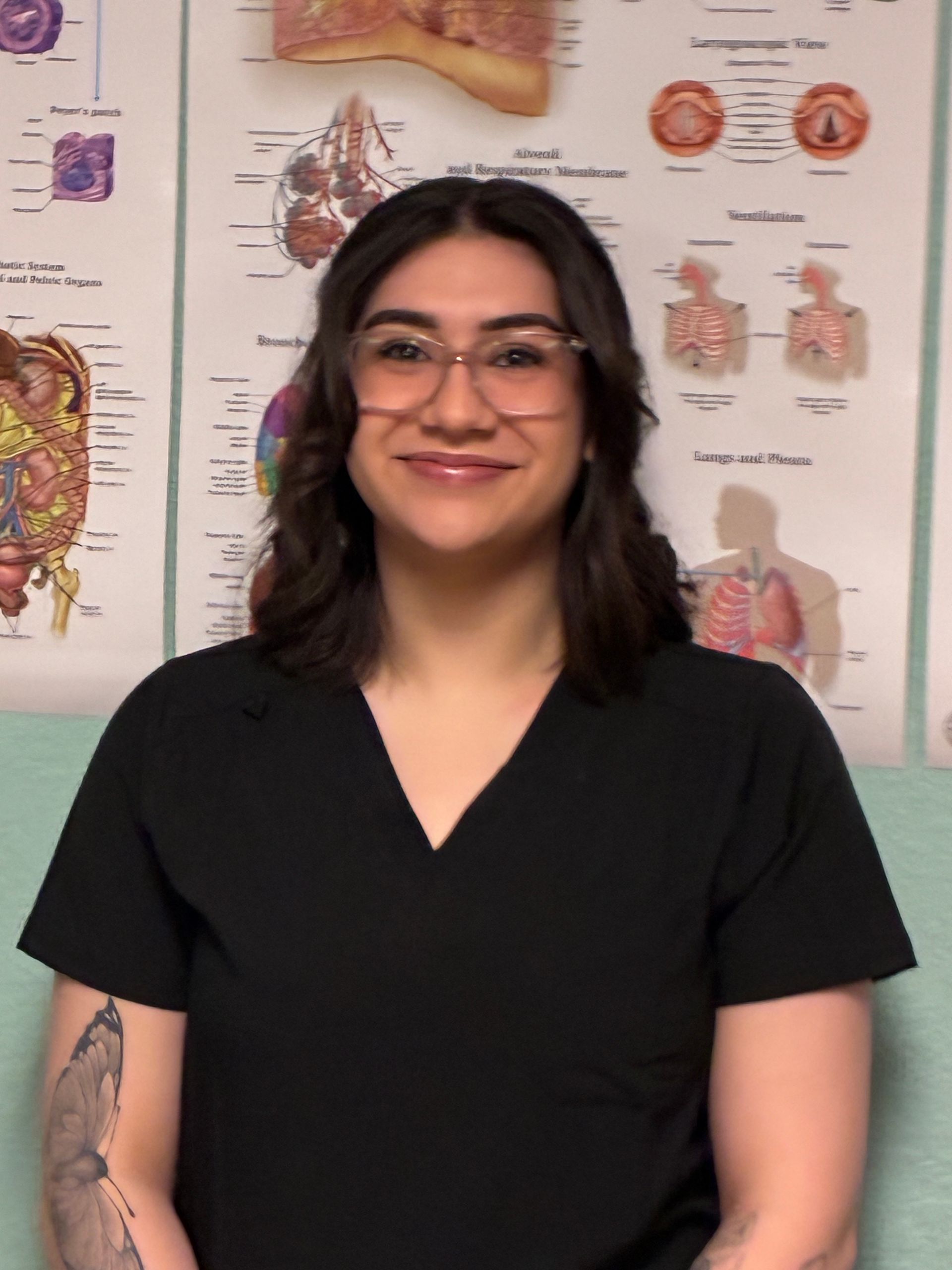 Smiling person in black scrubs standing in front of anatomical posters