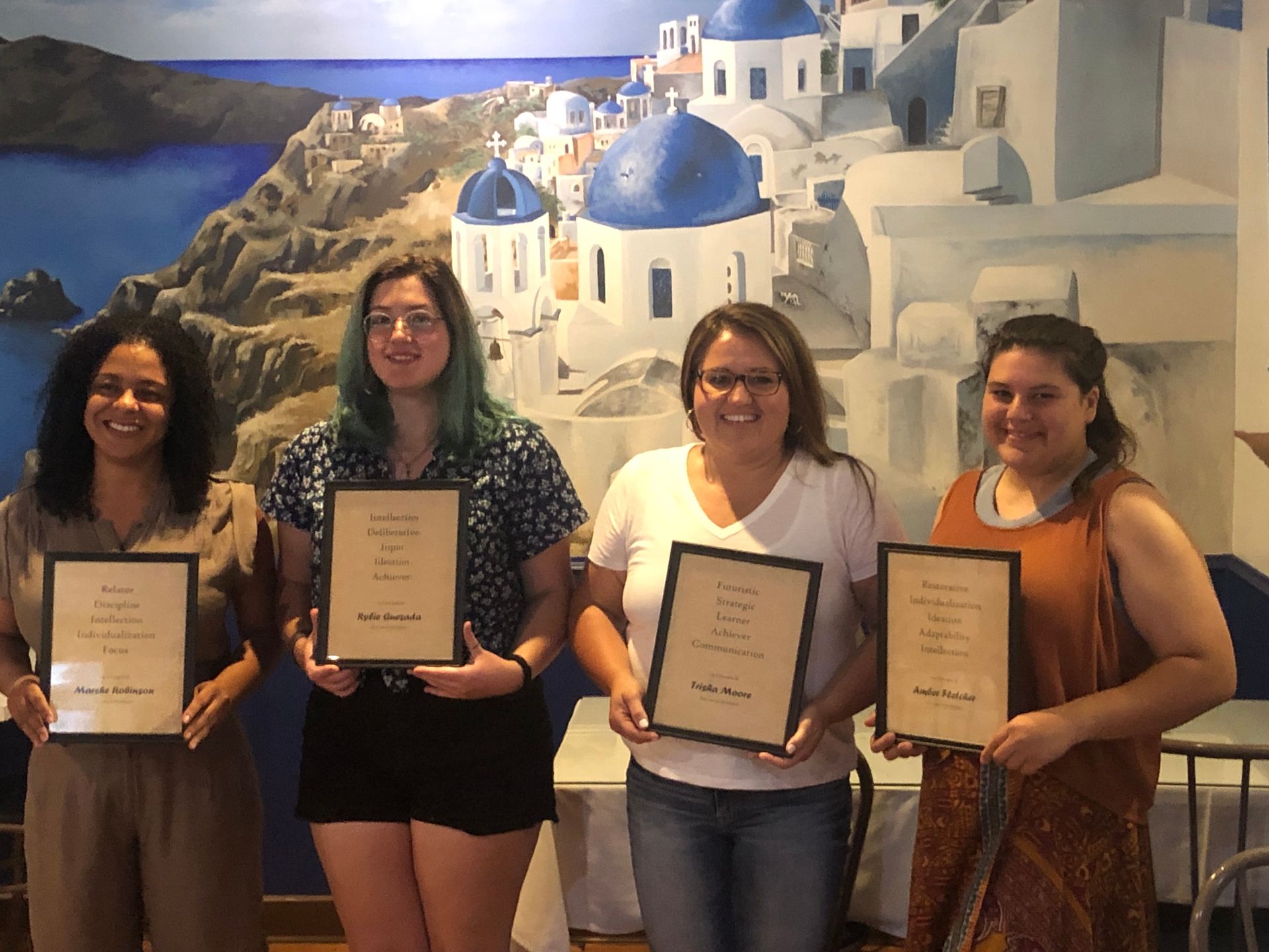 Four people holding framed certificates, smiling in front of a blue-and-white coastal mural.