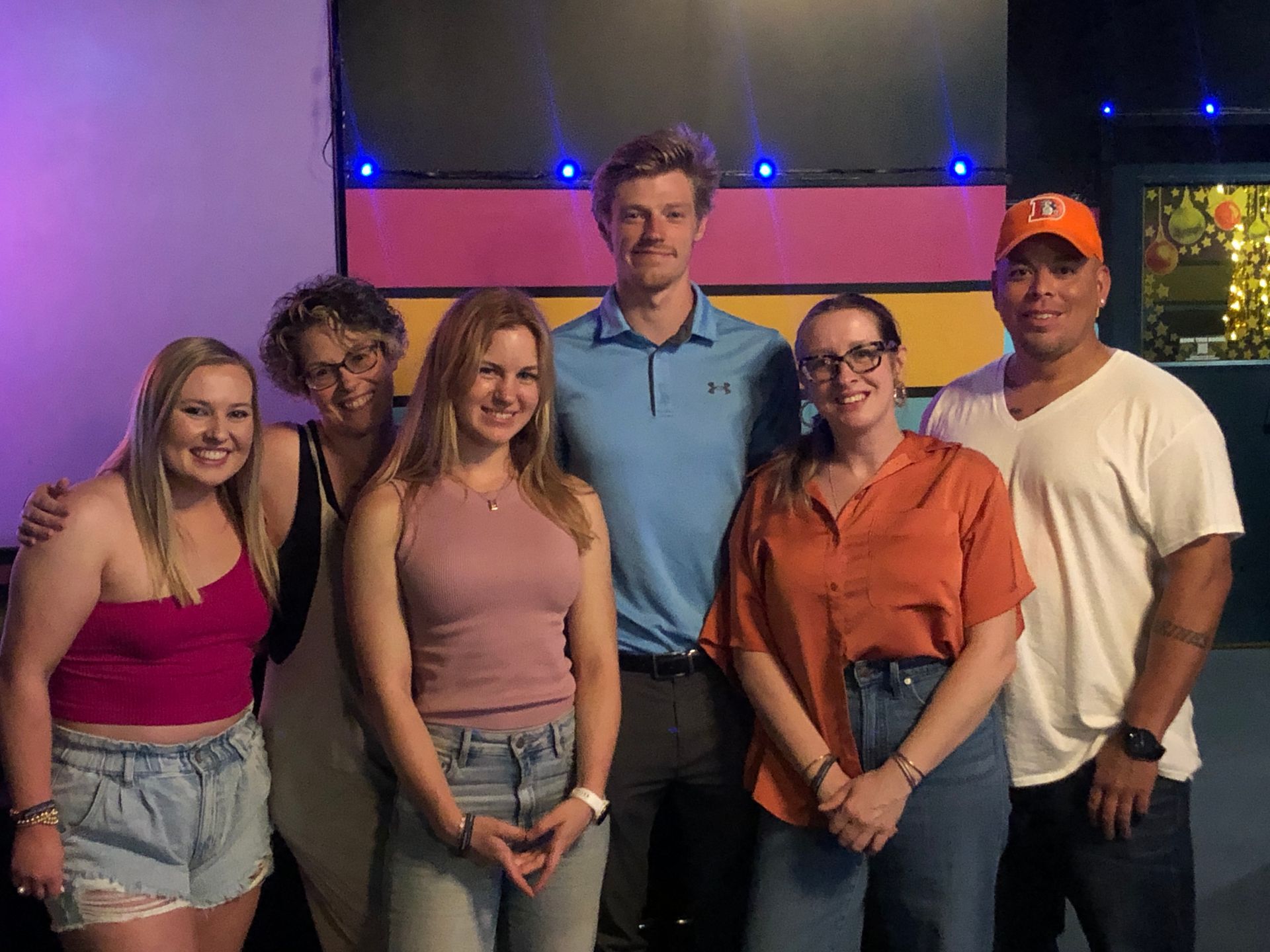 Six people posing indoors in front of purple-lit panels, smiling at the camera.