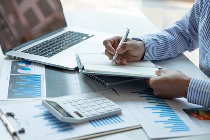 Person taking notes beside laptop, calculator, and blue financial charts on a desk