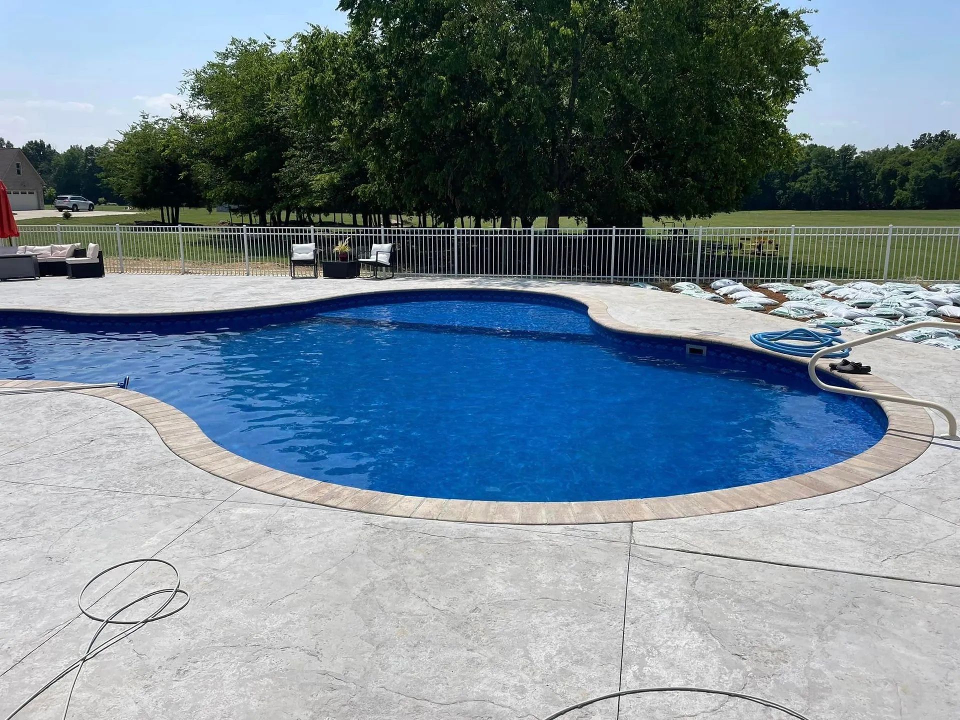 A kidney-shaped swimming pool filled with blue water. Gray concrete surrounds the pool.  White fence and trees in the background.