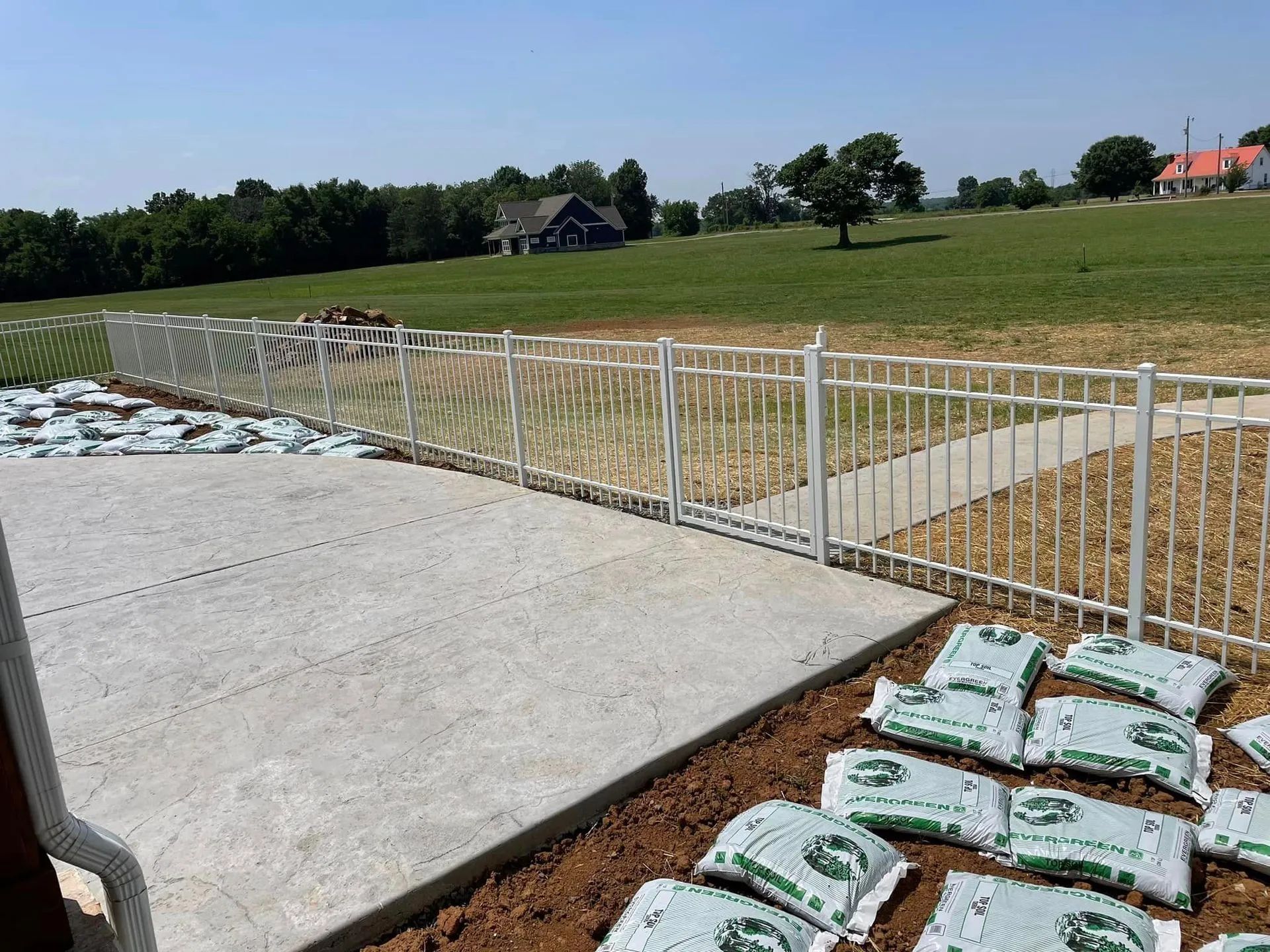 White metal fence on a concrete patio, with bags of planting material. In a grassy field, a house is visible.