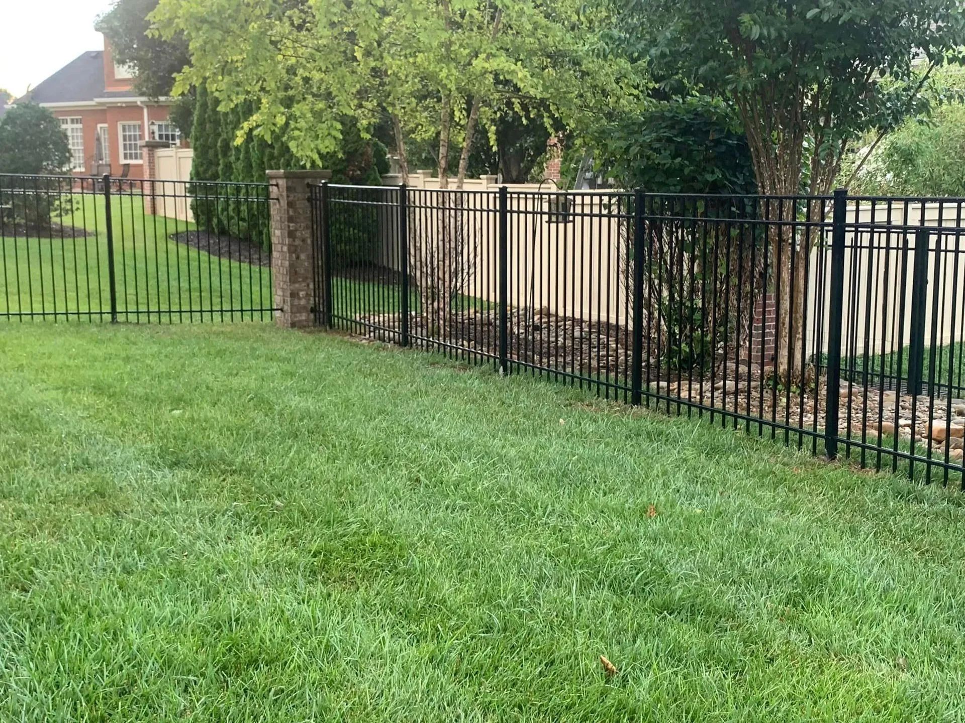 Black metal fence encloses a green lawn, near trees and a brick house.