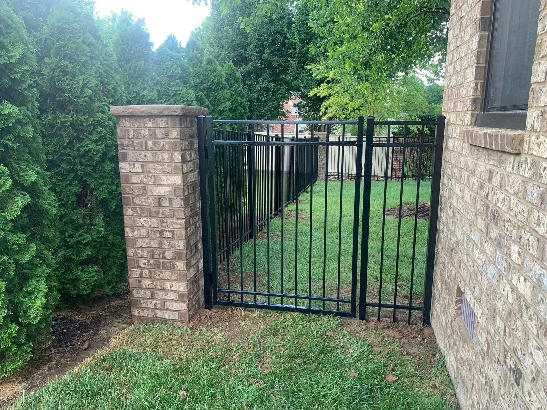 Black metal fence and gate between a brick column and house, grassy area.