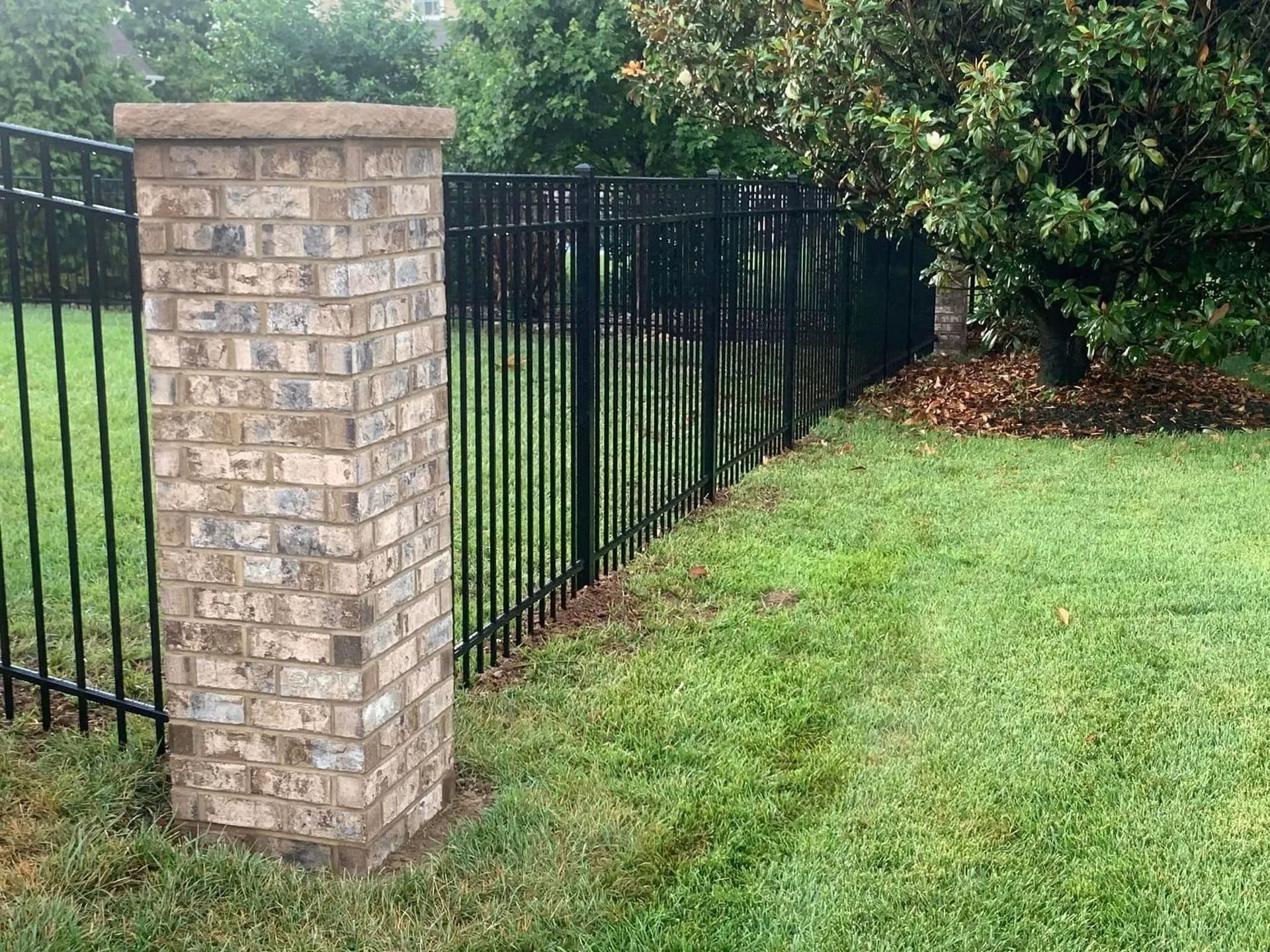 Brick pillar supporting a black metal fence in a grassy yard, next to trees.