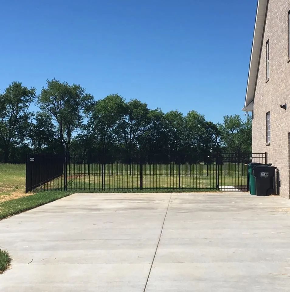 Black metal fence enclosing a grassy yard, concrete driveway, trees, and a brick house under a blue sky.