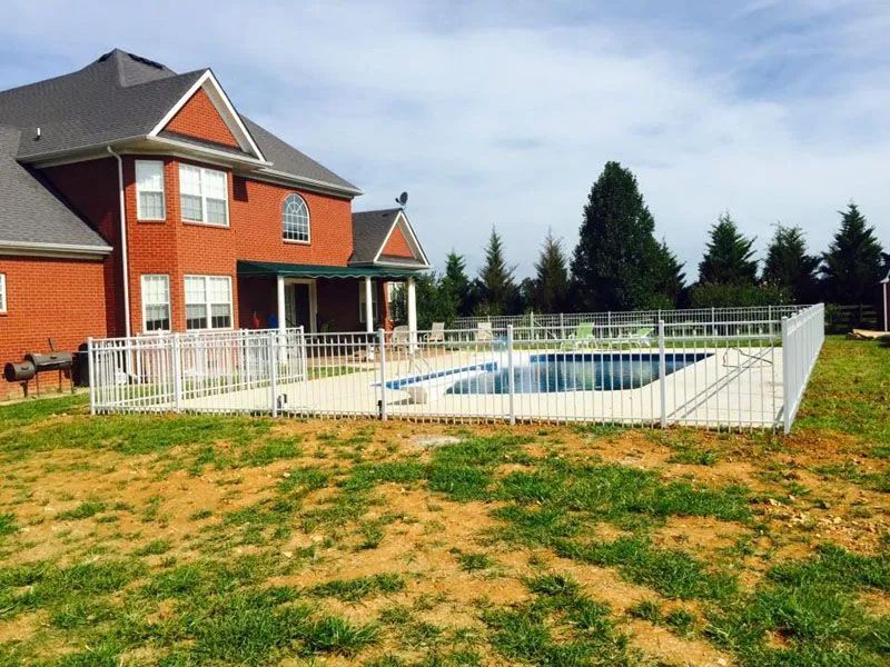 White fenced-in outdoor pool next to a red brick house with a green lawn.