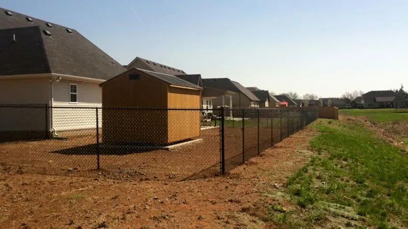 Chain link fence bordering residential properties and a grassy field. A shed sits within the fence line.