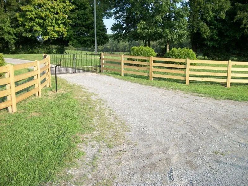 Gravel driveway leads to a gated entrance with a wooden fence on either side, grassy areas, and trees.