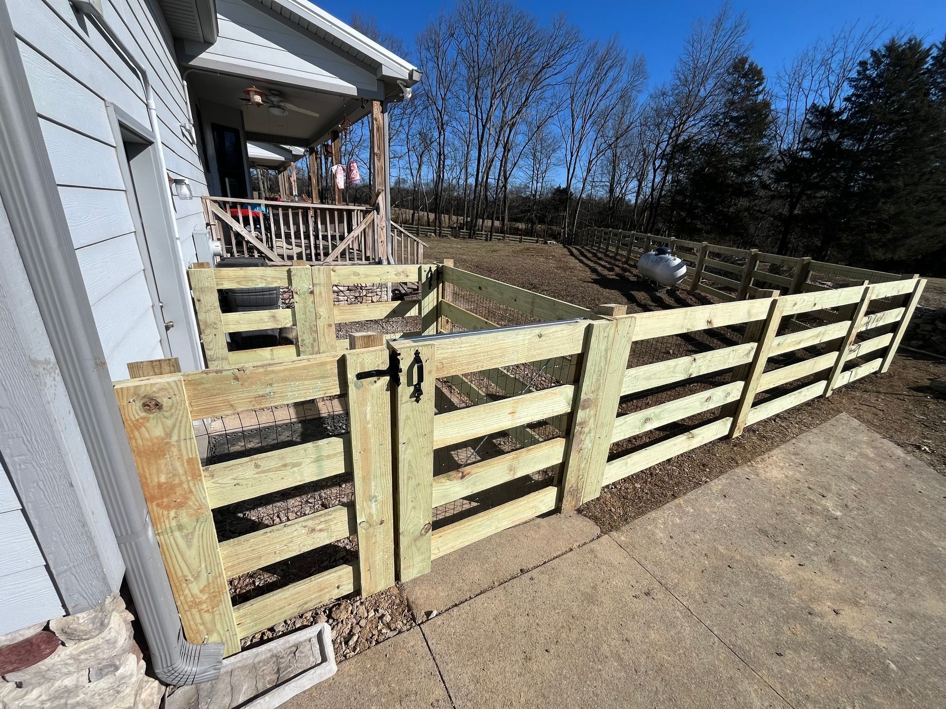 Wooden fence surrounding a concrete patio, attached to a house with a porch and trees in the background.
