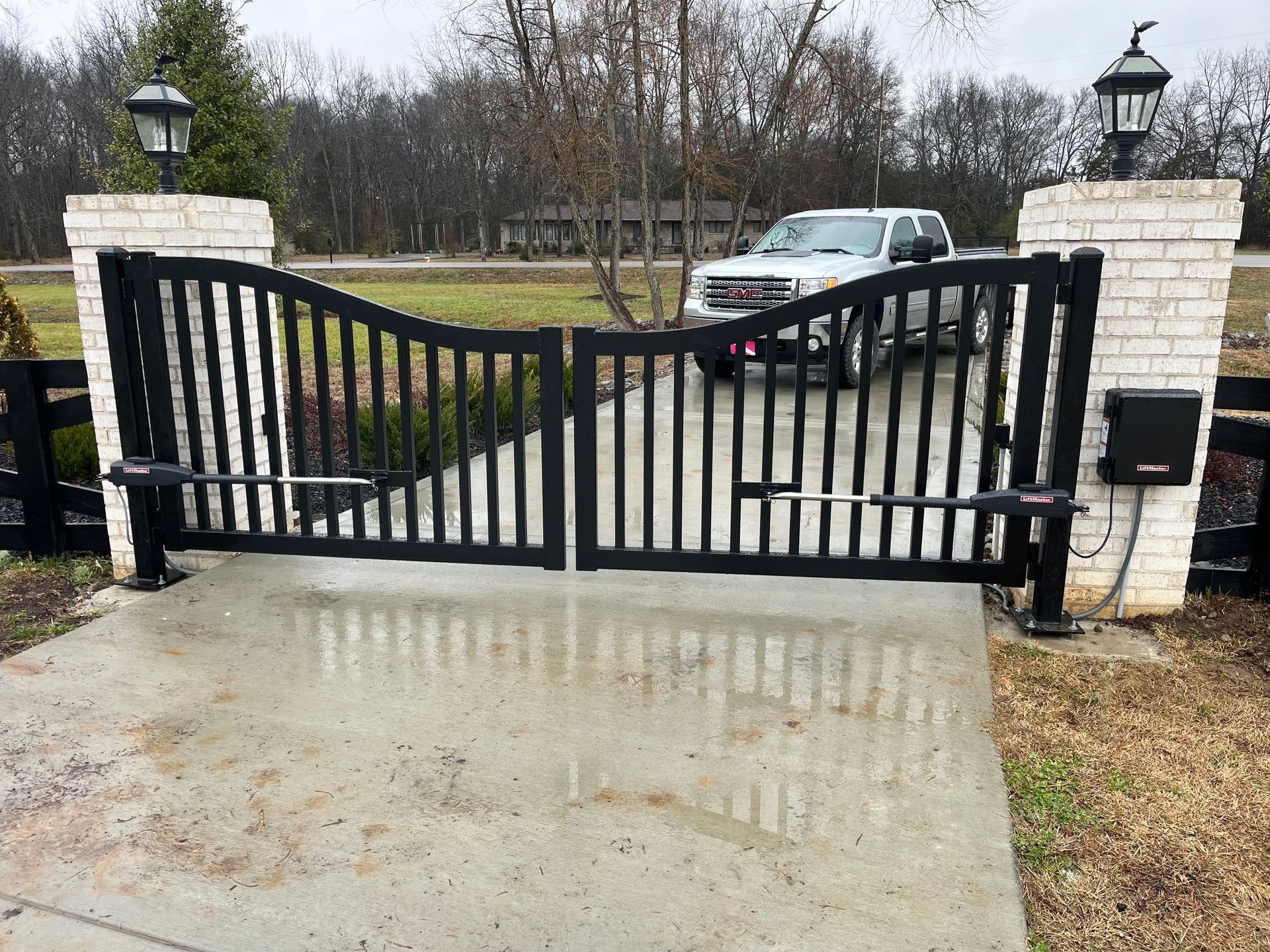 Black metal driveway gate, open, with a white truck parked in the driveway; brick columns with lanterns.