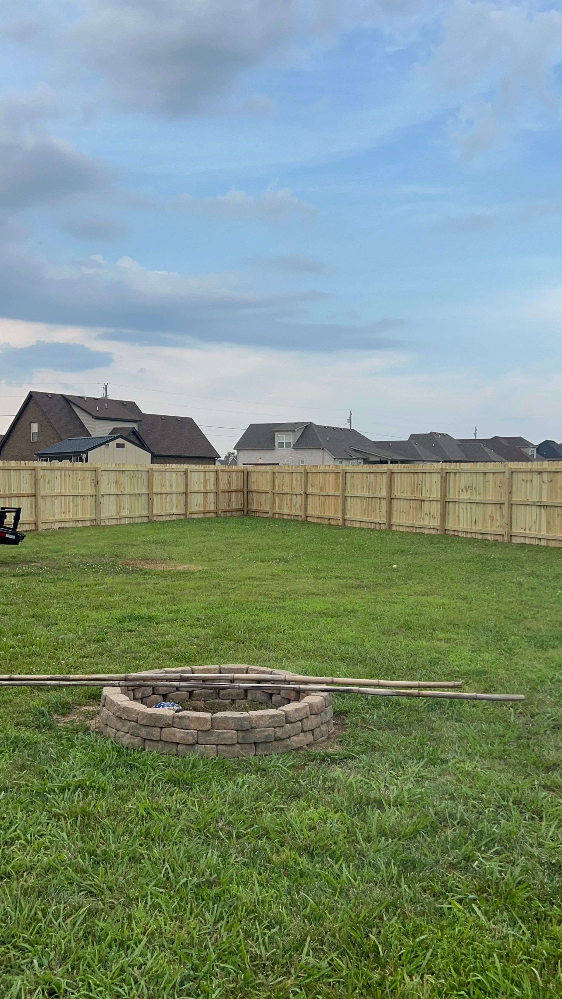 Backyard with a wooden fence, fire pit, and houses under a cloudy sky.