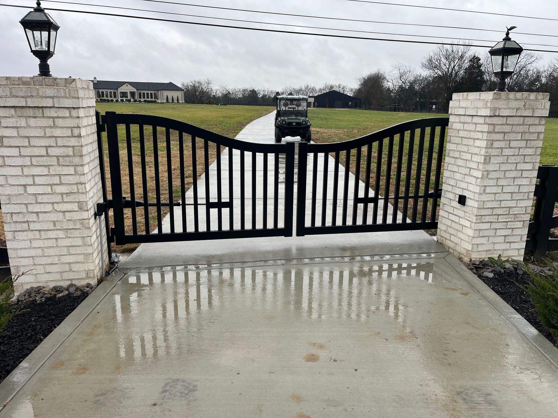 Black metal gate between brick pillars on a concrete driveway, leading to a distant house and vehicle.