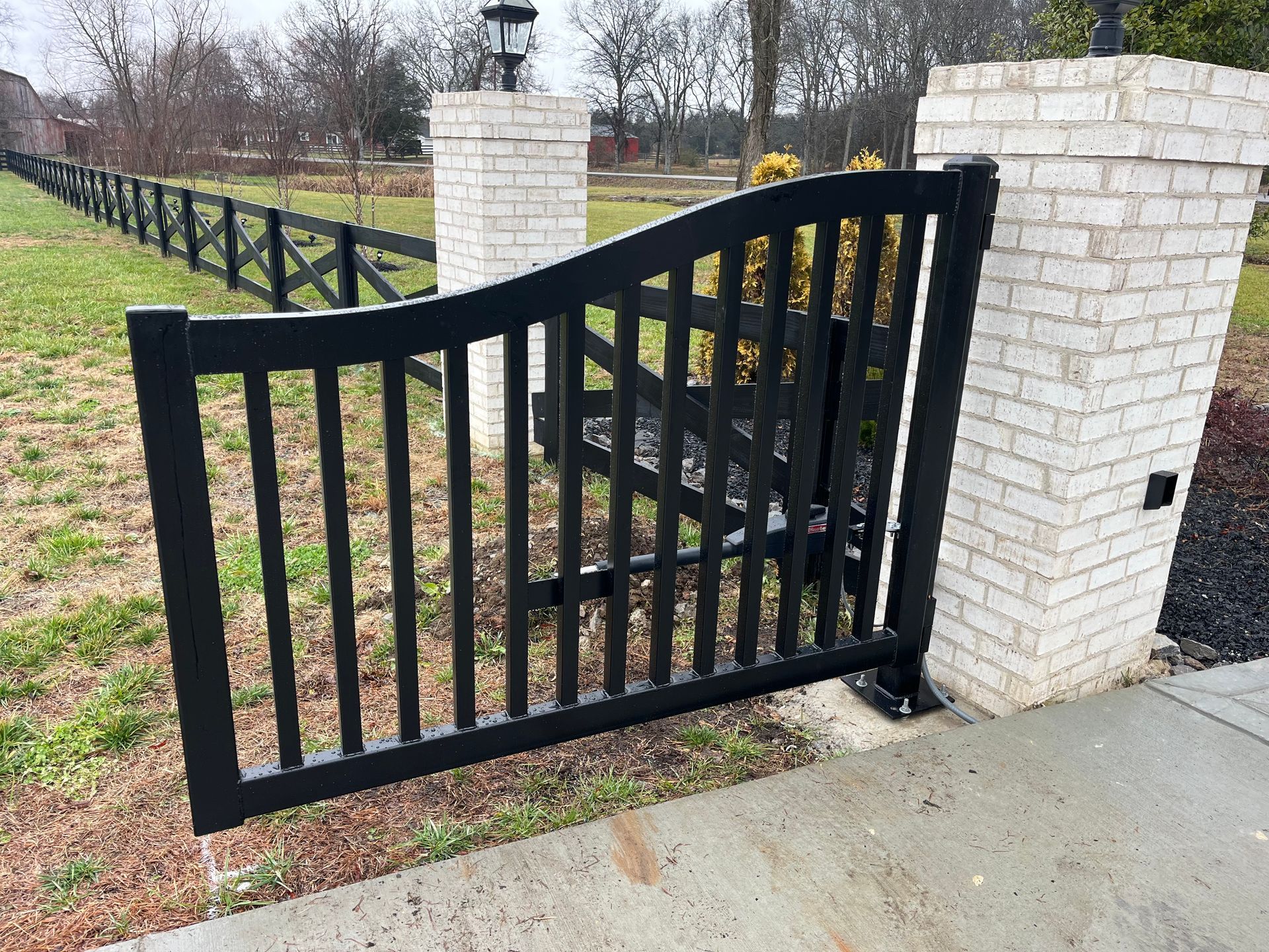Black gate in a brick pillar opening onto a grassy area with a black fence.