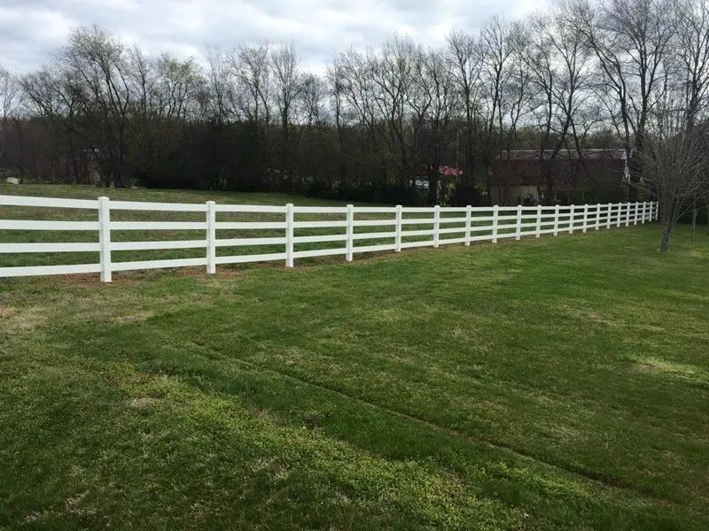 White, three-rail fence in a grassy yard, with a treeline in the background under a cloudy sky.
