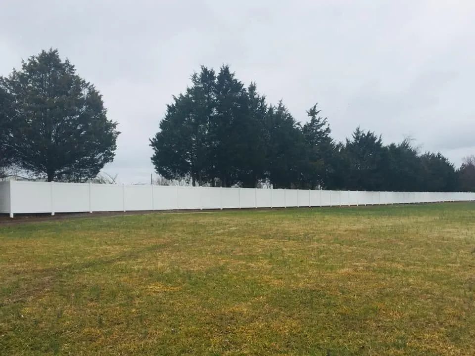 White vinyl fence in a grassy field, trees in background, overcast sky.
