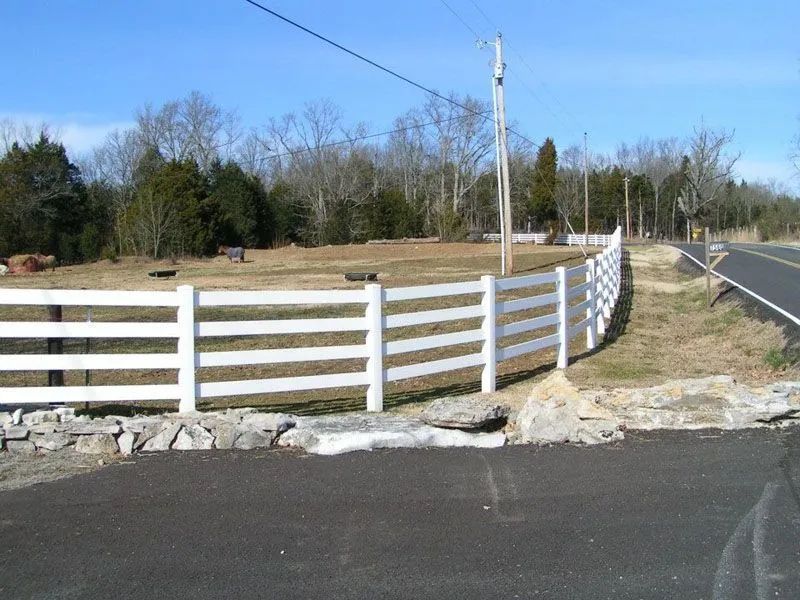 White fence borders a grassy area next to a paved road, under a clear sky.
